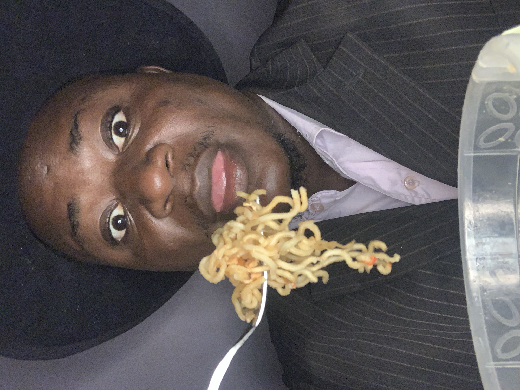 In Akwanga, Nigeria, a close-up, front-facing image captures an adult male with dark skin and a dark hat. His eyes are wide open, pupils dilated, and he looks directly at the camera. His mouth is open with his tongue extended. He wears a dark pinstripe suit jacket over a light-colored collared shirt. A silver fork is positioned directly in front of his mouth, holding a substantial portion of light-colored, cooked instant noodles with visible red specks of seasoning. Below the noodles, the rim of a clear plastic food container is visible. The background is uniformly dark.