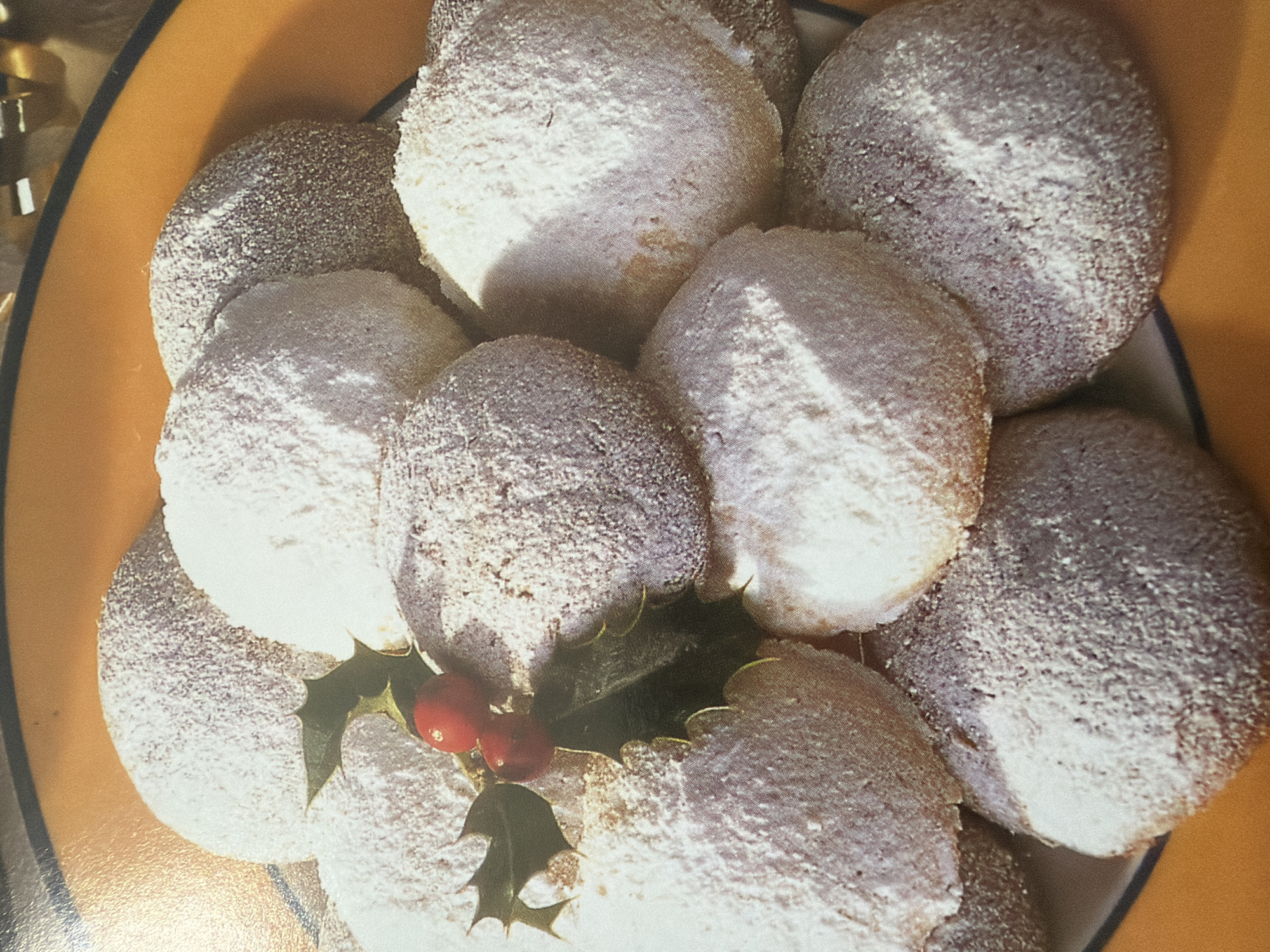 A close-up overhead shot shows a plate of Christmas cookies sprinkled with powdered sugar. The cookies are round and dome-shaped, with some having a darker, possibly chocolate, interior visible where the powdered sugar is thin. A sprig of holly with two red berries is nestled among the cookies. The cookies are arranged in a slightly overlapping, rustic heap on what appears to be a ceramic plate with a textured orange rim. The lighting creates shadows and highlights that emphasize the powdery texture of the cookies. The background is out of focus, with hints of a yellow or orange surface. There are no people or identifiable city elements in the image.