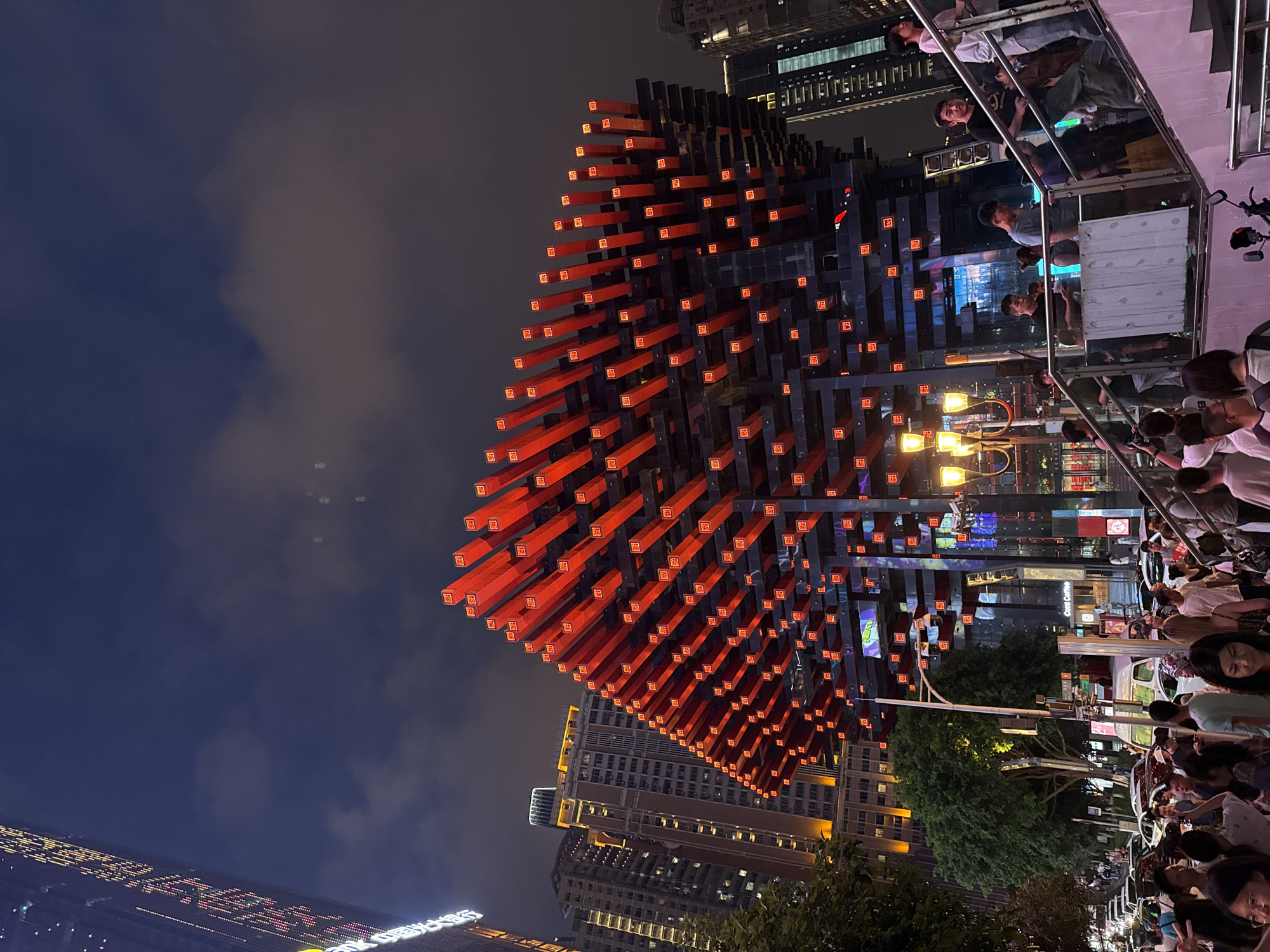 A night exterior shot captures the striking architecture of the Hongyadong Cave Dwelling in Yuzhong District, China. The building, illuminated with a warm glow, features a distinctive, layered design with numerous red, illuminated rectangular elements protruding from its facade. The surrounding area is a bustling urban environment, with several high-rise buildings visible in the background, their lights reflecting against the dark night sky. A crowd of people is present, with some ascending a staircase on the right side of the frame, while others are walking on a street level in the background, which is lined with cars and a green tree. Streetlights and building lights create a vibrant, nocturnal city scene.