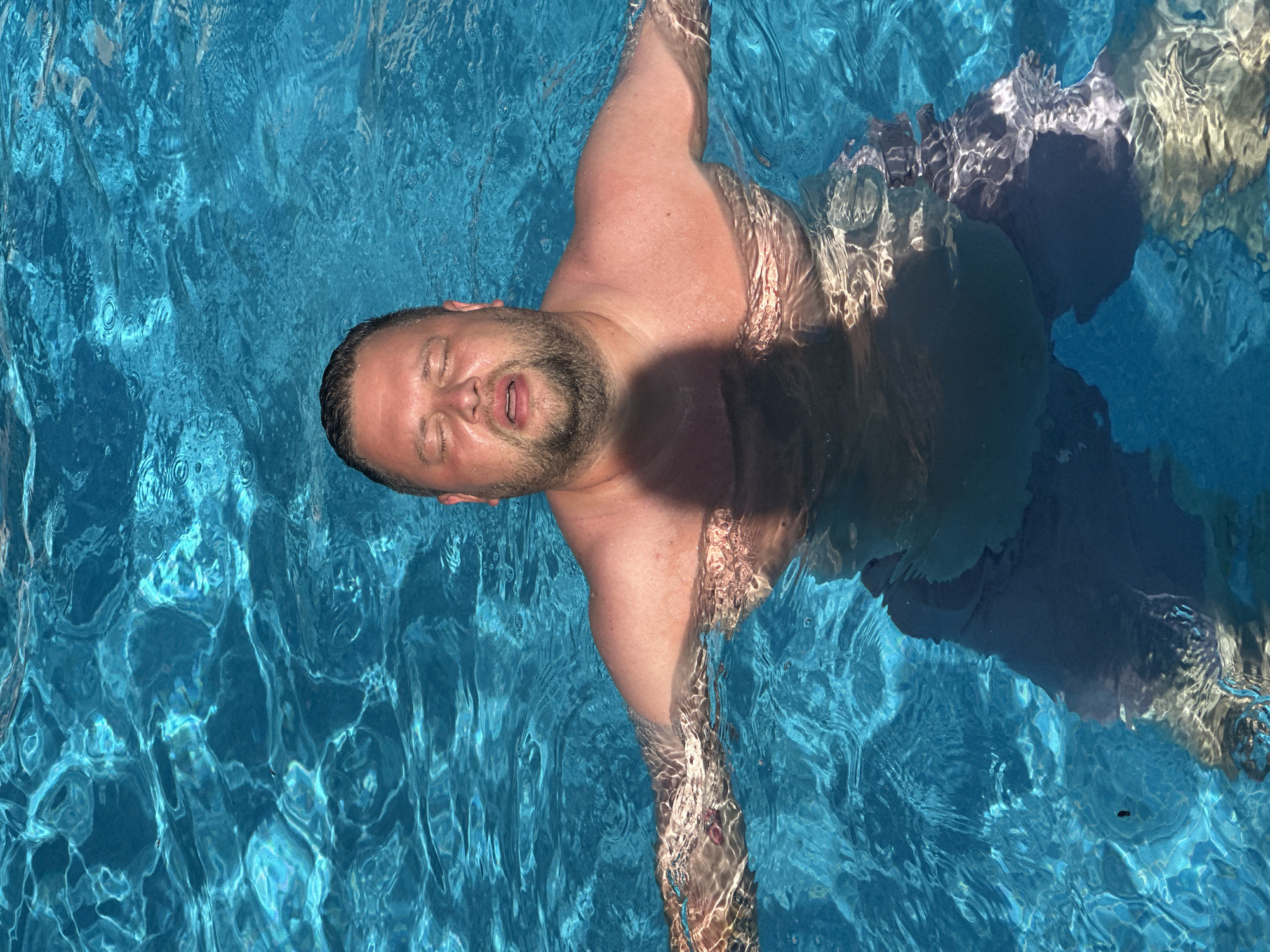 In Sant Feliu de Guíxols, Spain, a person is floating in a bright blue swimming pool. The person is submerged up to their chest, with arms outstretched to the sides and eyes closed. The water's surface is rippled, creating patterns of light and shadow on the person's skin. The person is wearing dark colored shorts. The overall impression is one of relaxation and enjoyment of the water on a sunny day.