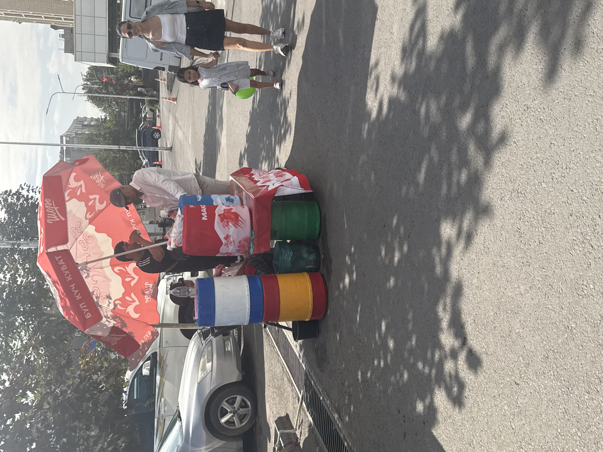 This image captures a street scene in Bishkek City, Kyrgyzstan, on a sunny day. In the foreground, a vendor is operating a small cart under a large red umbrella adorned with white patterns and Cyrillic text. The umbrella also features the logo "Шоро" (Shoro), a popular Kyrgyz beverage brand.
The vendor, a man wearing a dark t-shirt and a baseball cap, is seen drinking from a cup. Beside him, another man in a light-colored shirt and a cap is attending to the cart, which displays several colorful cylindrical containers stacked on top of each other, likely for drinks or ice. Some of these containers are red, yellow, blue, and white. There are also green bins and a red cooler with a white cloth draped over it.
In the background, a woman and a young girl are walking hand-in-hand. The woman wears a white tank top, a greyish-blue jacket, and a dark skirt, along with white sneakers. The girl is dressed in a light-colored dress and also wears sneakers, carrying a bright green spherical object in her left hand. They are walking away from the vendor.
Various vehicles are visible in the midground, including a silver car on the left and a white van and other cars further down the street. The setting appears to be a street or sidewalk with trees casting shadows on the asphalt. Buildings are visible in the background, suggesting an urban environment. A drainage grate runs along the edge of the sidewalk in the lower left corner. The overall impression is of a typical day with commercial activity and pedestrian movement in a city.