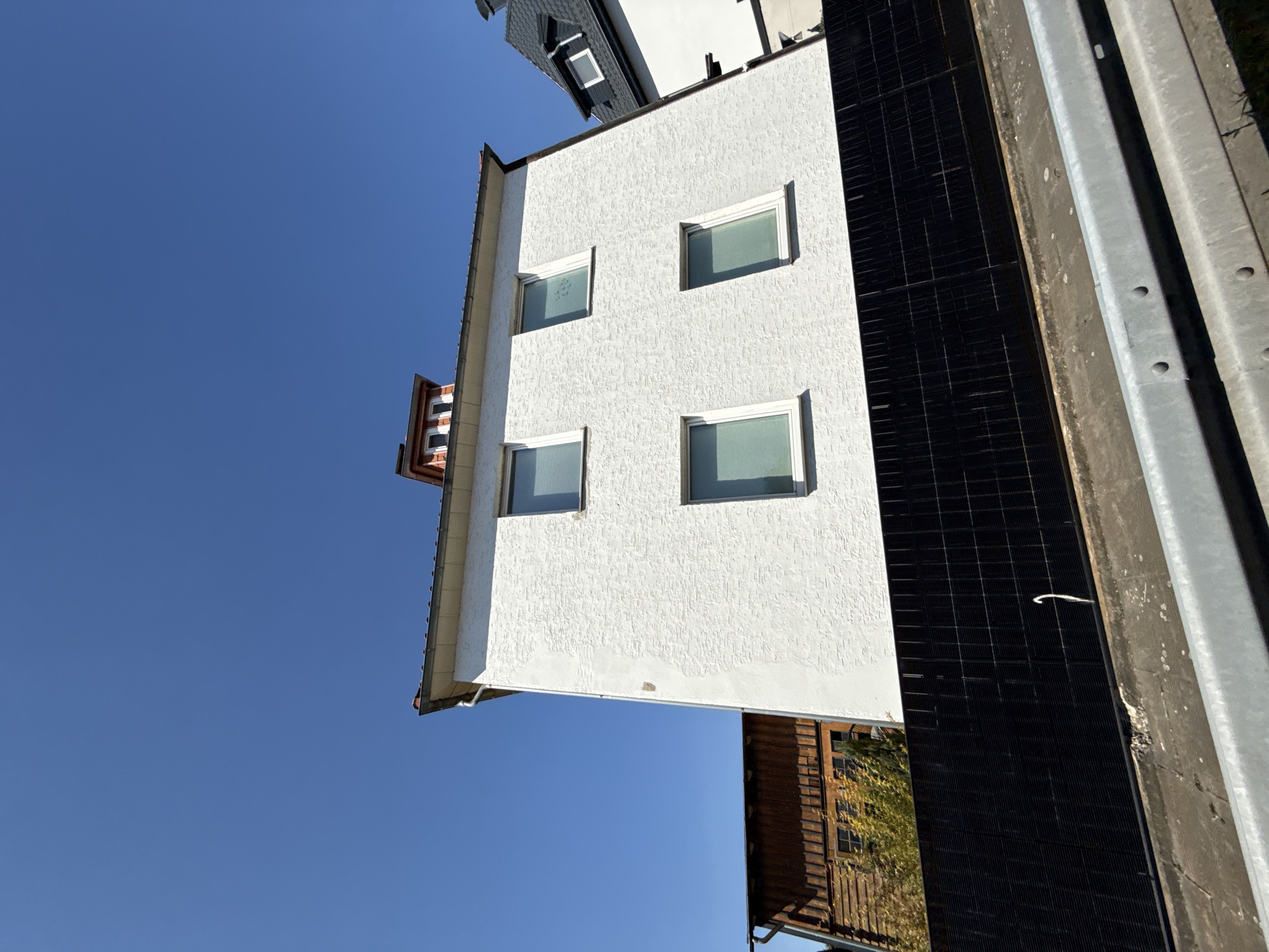 In Herzberg, Germany, a white building with a textured facade stands against a clear blue sky. The building features four rectangular windows on its visible side. A dormer window structure with a red and white striped pattern sits atop the roof. To the left of the white building, a darker wooden structure with visible windows is partially obscured by foliage. A black metal fence runs horizontally across the lower portion of the image, separating the buildings from the foreground. The foreground shows a concrete surface with a visible metal railing.
