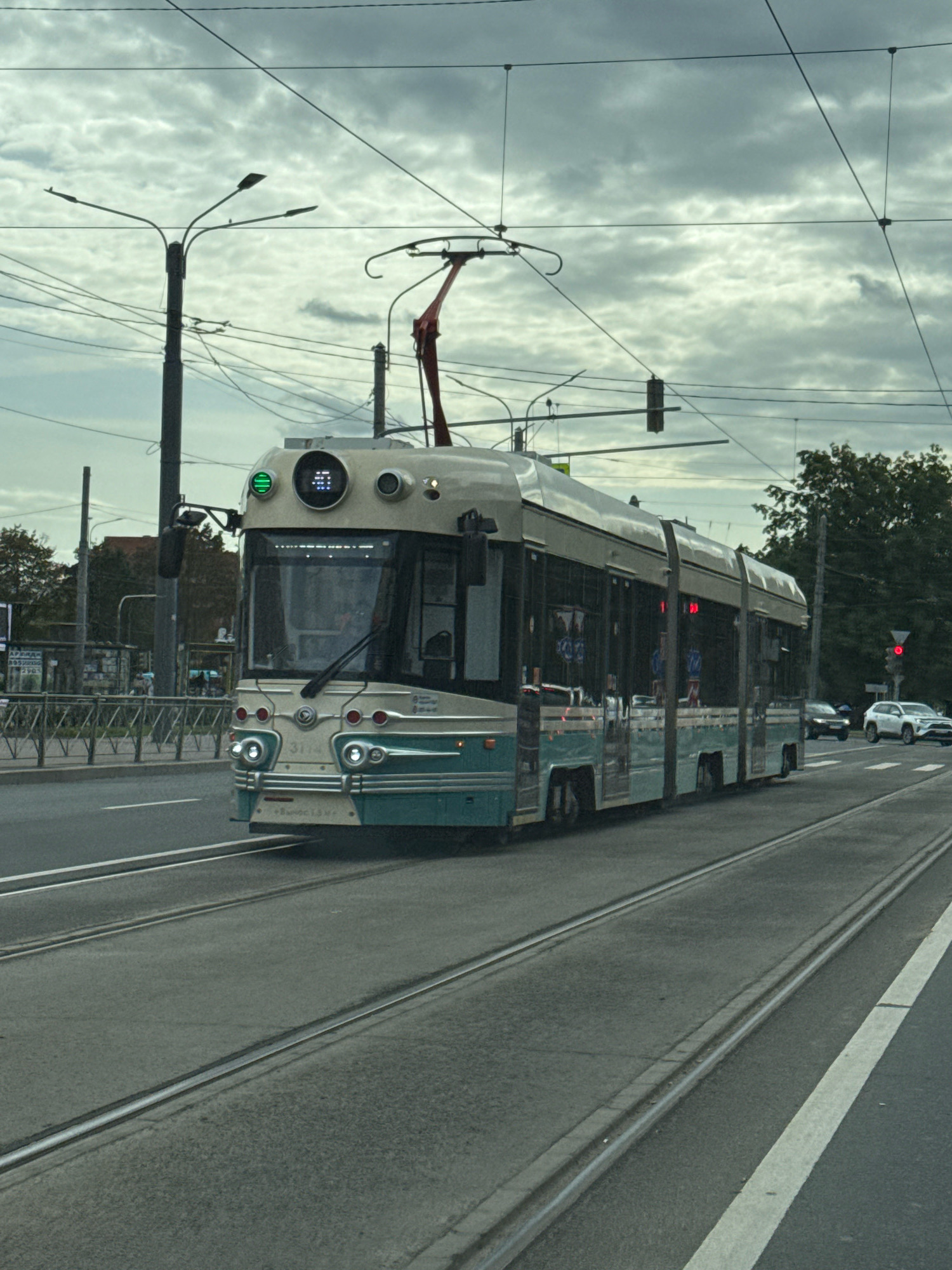 This is a medium-distance, eye-level, full-frame shot of a tram moving on tracks in Saint Petersburg, Russia, on a cloudy day. The tram is a long, articulated vehicle, painted in a cream and teal color scheme. It is captured in motion, traveling from left to right in the frame. The front of the tram is prominently visible, featuring a digital display with green lights, round headlights, and a smaller auxiliary light. The number "3114" is visible on the front of the tram. The tram is surrounded by overhead electrical wires and poles, typical of urban tram systems. In the background, there are trees, buildings, and other vehicles, including a white SUV and a dark car, on the right side of the road. A traffic light is visible in the distance on the right, showing a red signal. The road is paved, with distinct tram tracks embedded in it. The sky is overcast with a dramatic cloud formation. The image appears to be taken from another vehicle, likely also a tram or car, moving alongside the subject tram.