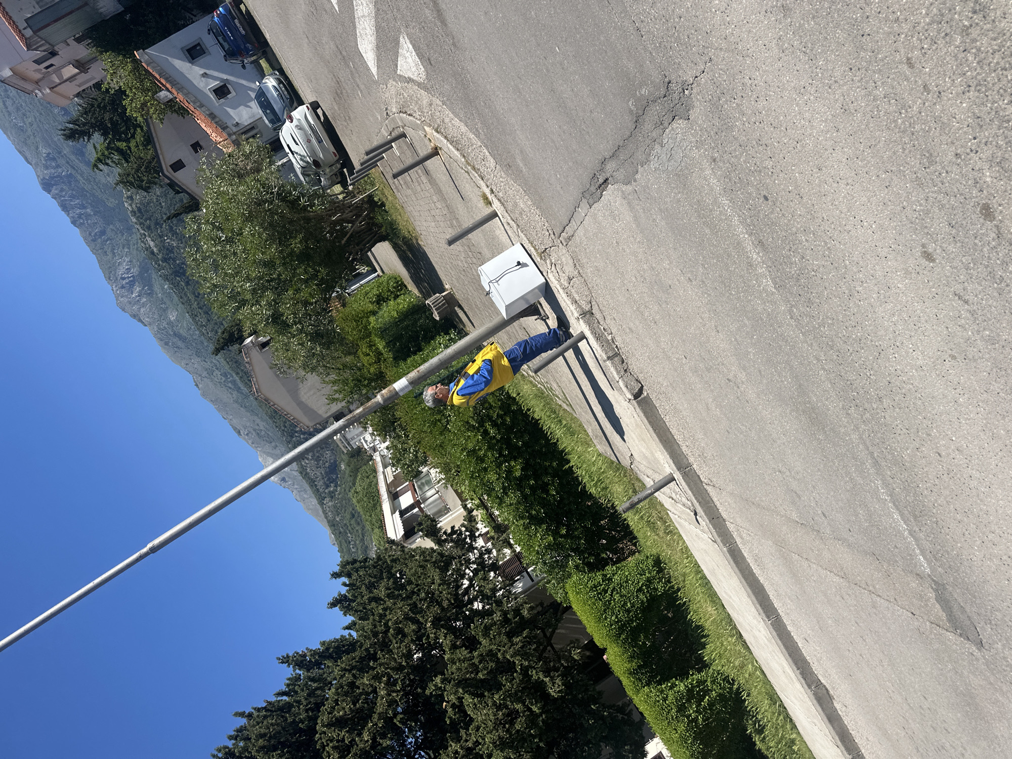 The image captures a scene in Budva, Montenegro, near the Jadranski put road and a road-side electrical box. A man wearing a blue and yellow jacket stands near a gray metal pole. The surrounding environment includes trees, grass, a concrete curb, and an asphalt road. In the background, residential buildings and a mountain can be seen under a clear blue sky. Several cars are parked along the road further up the road.