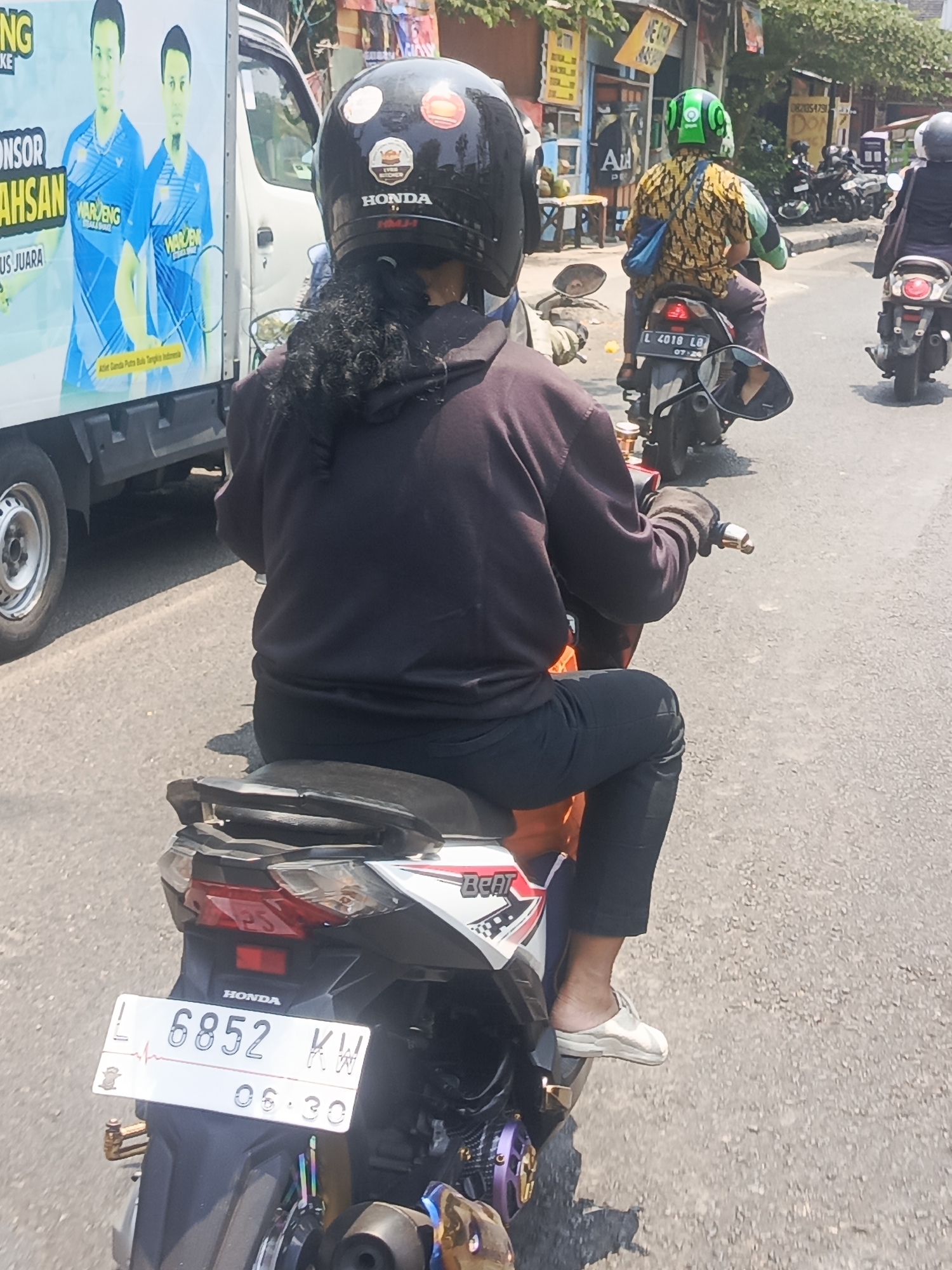 The image depicts a street scene in Surabaya, Indonesia, captured from the perspective of someone riding a motorcycle. In the foreground, a person wearing a black hoodie, black pants, and a black helmet with several stickers, including a Honda logo, is riding a white and orange motorcycle. Their curly dark hair is visible beneath the helmet. The motorcycle has a license plate that reads "L 6852 KW" and "06.30". The individual is wearing light-colored slip-on shoes.
To the left of the motorcycle, a white truck with a large advertisement on its side is partially visible. The advertisement features two male figures in blue athletic wear and the text "SPONSOR SAHSAN" and "US JUARA".
Further down the street, other motorcycles are visible, including one with a rider wearing a yellow and green patterned shirt and a green helmet. The environment appears to be a moderately busy urban street with buildings and shops lining the roadside. The lighting suggests it is daytime, with bright sunlight casting shadows. The road surface is asphalt.