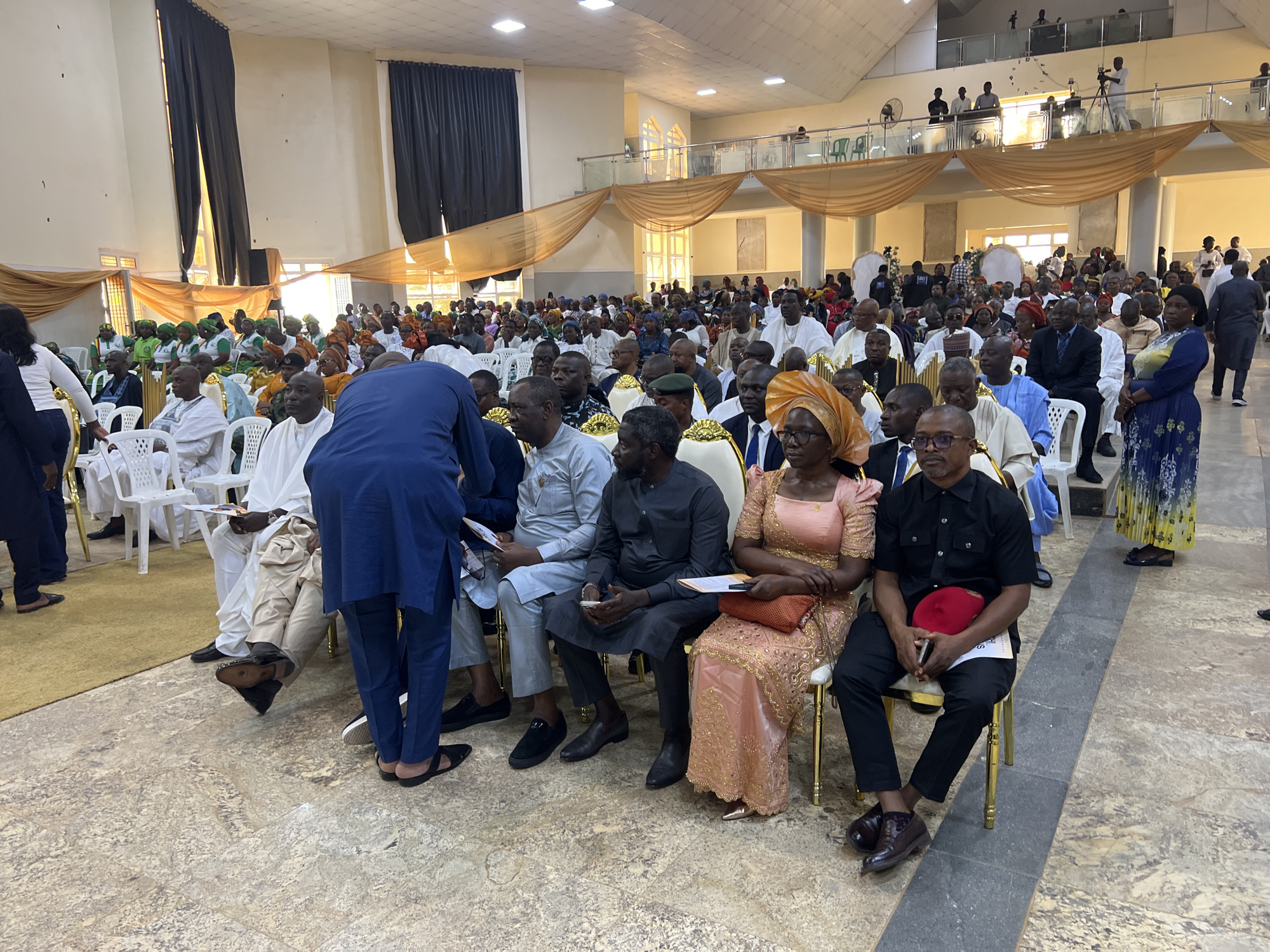 A large group of people are seated in a hall, likely for an event or conference, in Kabong, Nigeria. The hall is decorated with draped fabric in gold and black, and bright lighting illuminates the scene. Many attendees are dressed in traditional attire, with some men wearing agbadas and embroidered caps, while women are in colorful wrappers and headscarves.
In the foreground, a man in a dark blue suit stands with his back to the camera. To his left, a group of people are seated, including a woman in a pink and gold dress and a man holding a red cap. Further into the hall, more rows of chairs are filled with men and women, some engaging in conversation, while others appear to be listening attentively.
The flooring is a patterned tile, and in the background, a stage or raised platform is visible, with people standing and operating a camera on a tripod, suggesting the event is being recorded. The overall atmosphere appears formal and celebratory.