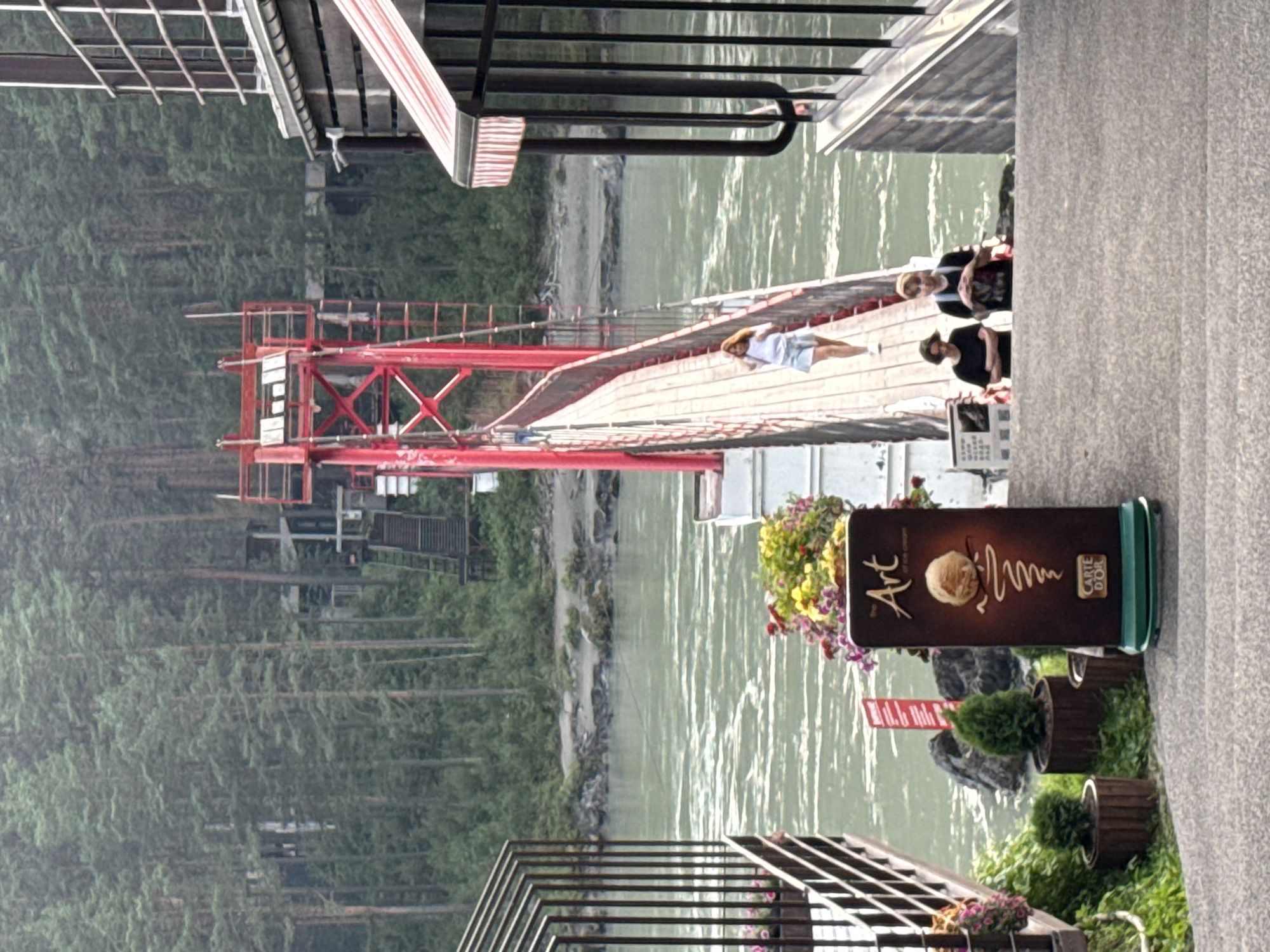 The image depicts a suspension bridge crossing a wide, turbulent river, likely the Katun River, near Barangol, Russia. The bridge's red metal structure and wooden walkway are prominent. On the bridge, a young woman with long, light brown hair, wearing a white top and denim shorts, walks away from the camera. In the foreground, on the right side of the image, two individuals are visible on a concrete path, one wearing a black t-shirt and the other a black t-shirt with a graphic print. To the left, a dark sign displays "The Art of ice cream" with an image of ice cream and "CARTE D'OR" at the bottom. In the background, a dense forest of tall pine trees lines the riverbank, with some structures partially visible amongst the trees. The overall atmosphere is one of a natural setting with recreational infrastructure.