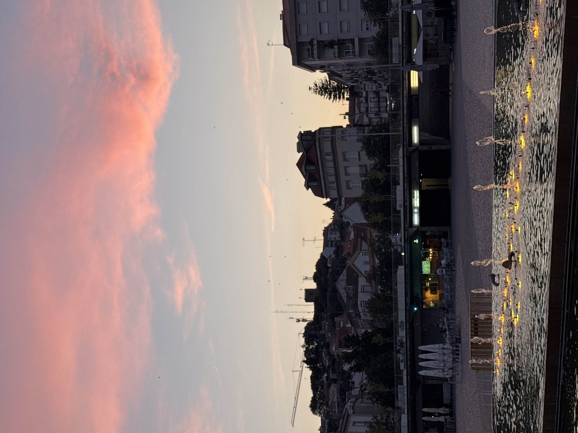 The image depicts a twilight scene in Castelo Branco, Portugal, focusing on a modern public space featuring a fountain and a cityscape in the background. The sky is painted with soft pink and orange hues, characteristic of a sunset, with a sliver of the moon visible near the horizon. Below the sky, the city of Castelo Branco unfolds with a mix of residential buildings and older structures, including a castle-like tower on a hill dotted with trees and antennas. A construction crane is also present on the hill, suggesting ongoing development.
In the foreground, a wide, shallow fountain is illuminated with warm yellow lights, creating a mesmerizing display as water jets shoot upwards, forming delicate arcs. The surface of the fountain water is textured with ripples. The area around the fountain is paved, and in the middle ground, a row of restaurants or cafes are visible with outdoor seating, some with umbrellas, and patrons can be seen in some of them. The overall atmosphere is calm and serene, capturing the transition from day to night in a Portuguese city.