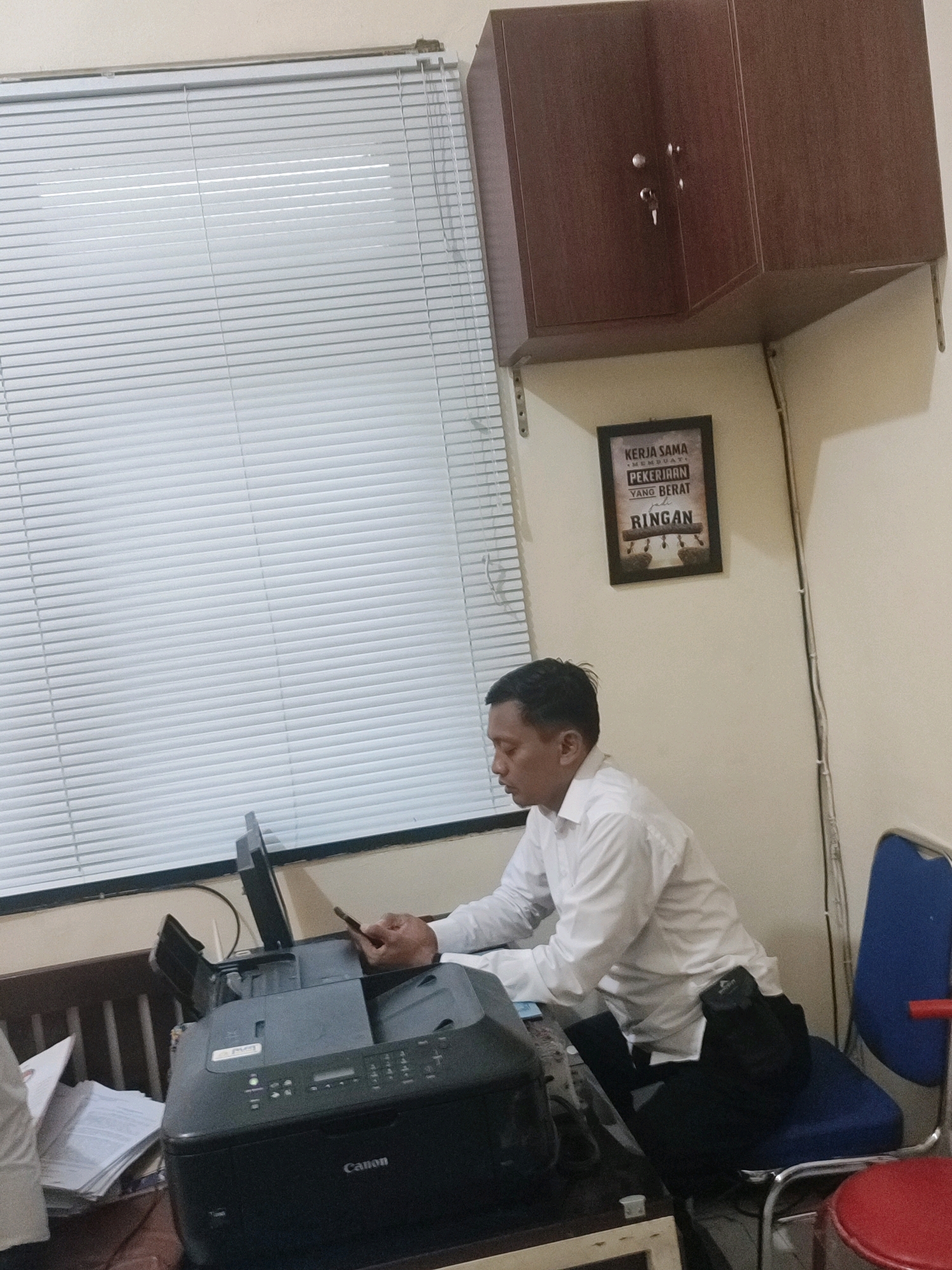 The image depicts an indoor scene, likely an office, in Ringinanom, Indonesia. A man wearing a white collared shirt and dark pants is seated at a desk, looking down at his mobile phone. In front of him is a black Canon printer, with its lid open. To the left of the printer, a stack of papers is visible. Behind the man, on the wall, hangs a framed motivational poster with text in Indonesian, which appears to read "KERJA SAMA MEMBUAT PEKERJAAN YANG BERAT RINGAN" (Working together makes heavy work light). Above the poster is a dark brown wooden cabinet mounted on the wall, featuring two silver knobs and a key. A white Venetian blind covers a window to the left of the frame. A blue chair with a silver frame is positioned to the right of the desk, and part of a red stool is visible in the foreground. The walls are painted a light, neutral color.