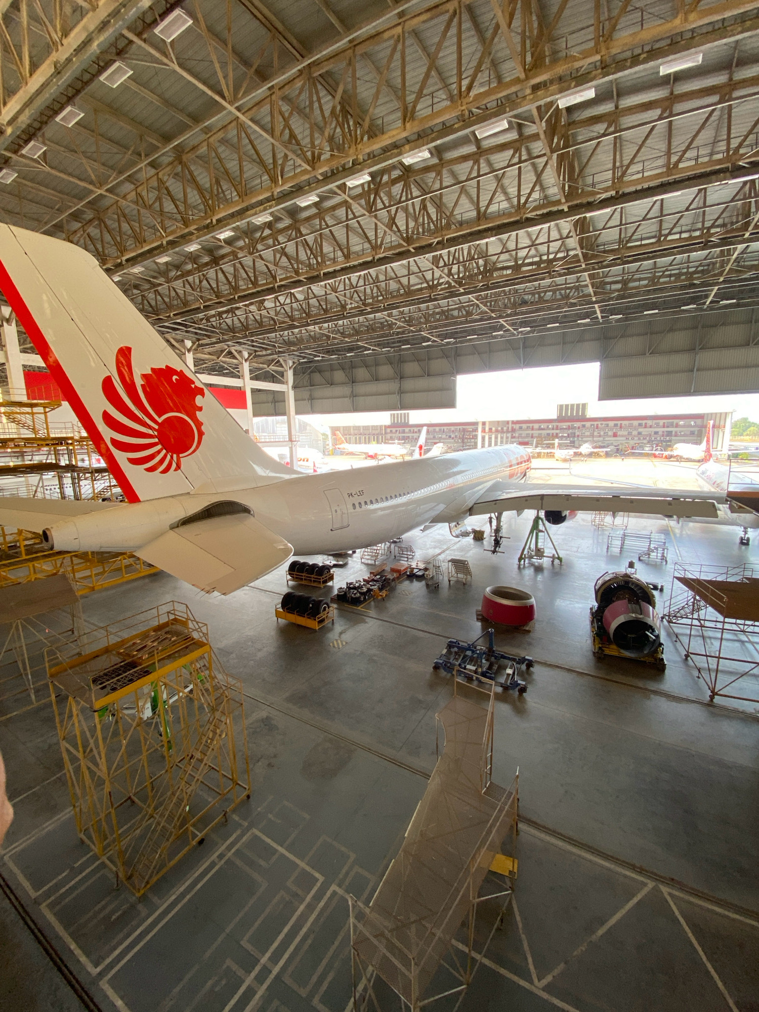 The image depicts the interior of a large aircraft hangar, likely located at Hang Nadim International Airport in Batam, Indonesia, given the presence of a Lion Air aircraft with the registration PK-LEF. The hangar is spacious, with a high, exposed metal truss ceiling and numerous fluorescent lights. A wide-open bay at the far end of the hangar reveals daylight and other aircraft parked outside.
The primary subject is a white airplane with a distinctive red Lion Air logo on its tail. The aircraft is partially disassembled or undergoing maintenance, as evidenced by the presence of various equipment around it. Directly beneath the tail and rear fuselage, there are several yellow scaffolding structures. In front of the aircraft's wing, another yellow scaffolding structure is positioned, along with a detached engine mounted on a yellow platform. Further towards the foreground, there are stacks of aircraft tires on yellow pallets, a red engine cowling on the floor, and a wheeled piece of agricultural-looking equipment. A temporary walkway or ramp extends from the foreground towards the aircraft.
The floor of the hangar is a smooth, grey concrete surface marked with white lines, possibly indicating parking or maintenance zones. One person can be observed near the front of the aircraft, appearing to be engaged in maintenance work. The overall atmosphere is one of industrial activity and aircraft repair.