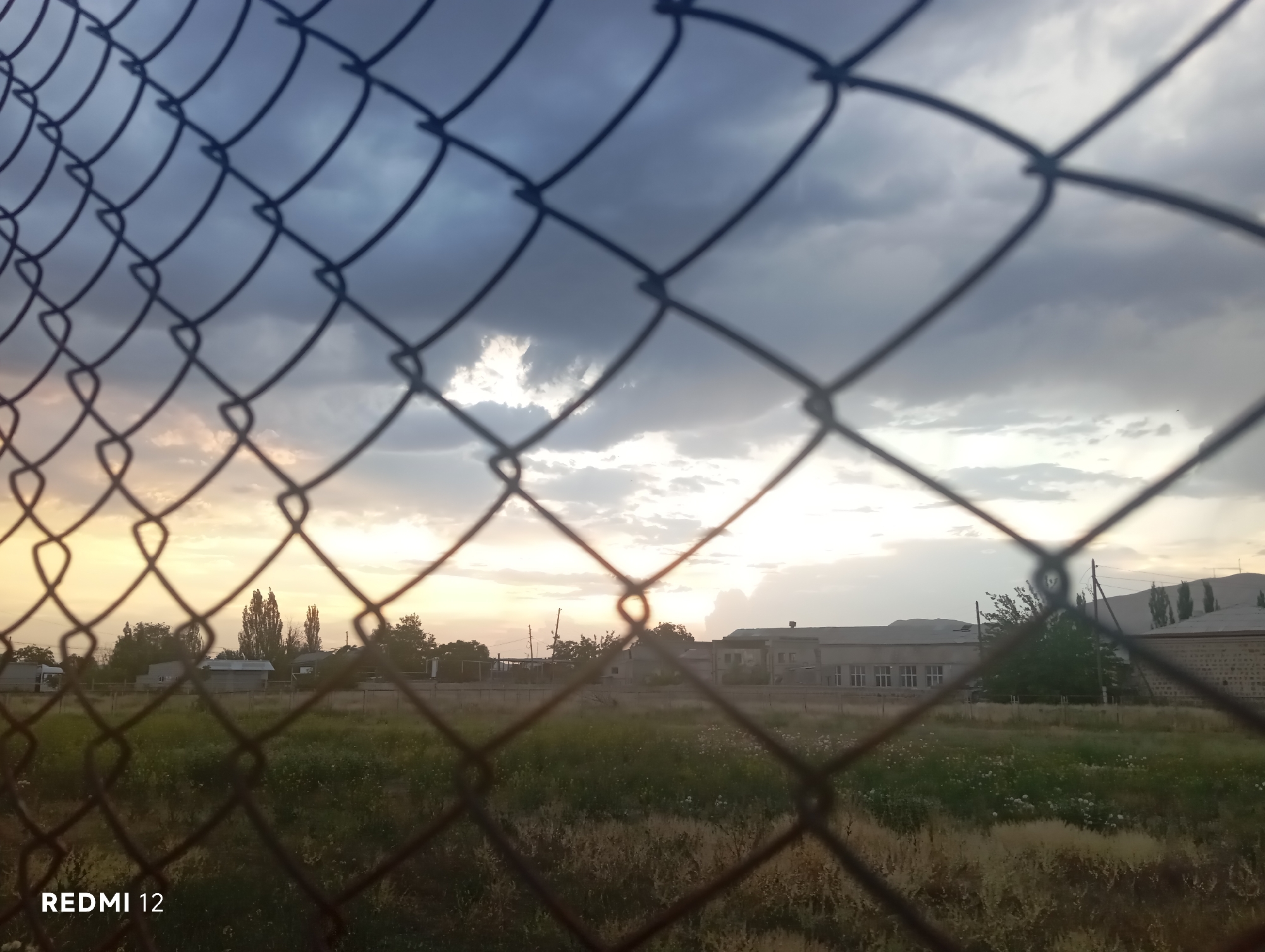 The image shows a cloudy sky at dusk, with the sun low on the horizon casting a warm glow. In the foreground, a chain-link fence is blurred, creating a diamond-shaped pattern that obscures the view. Beyond the fence, a field of dry grass stretches towards a line of buildings and trees. The buildings appear to be low-rise structures, possibly industrial or residential. The overall scene suggests a rural or semi-urban environment at the end of the day. There are no people visible in the image. The location is described as Unknown, Unknown.