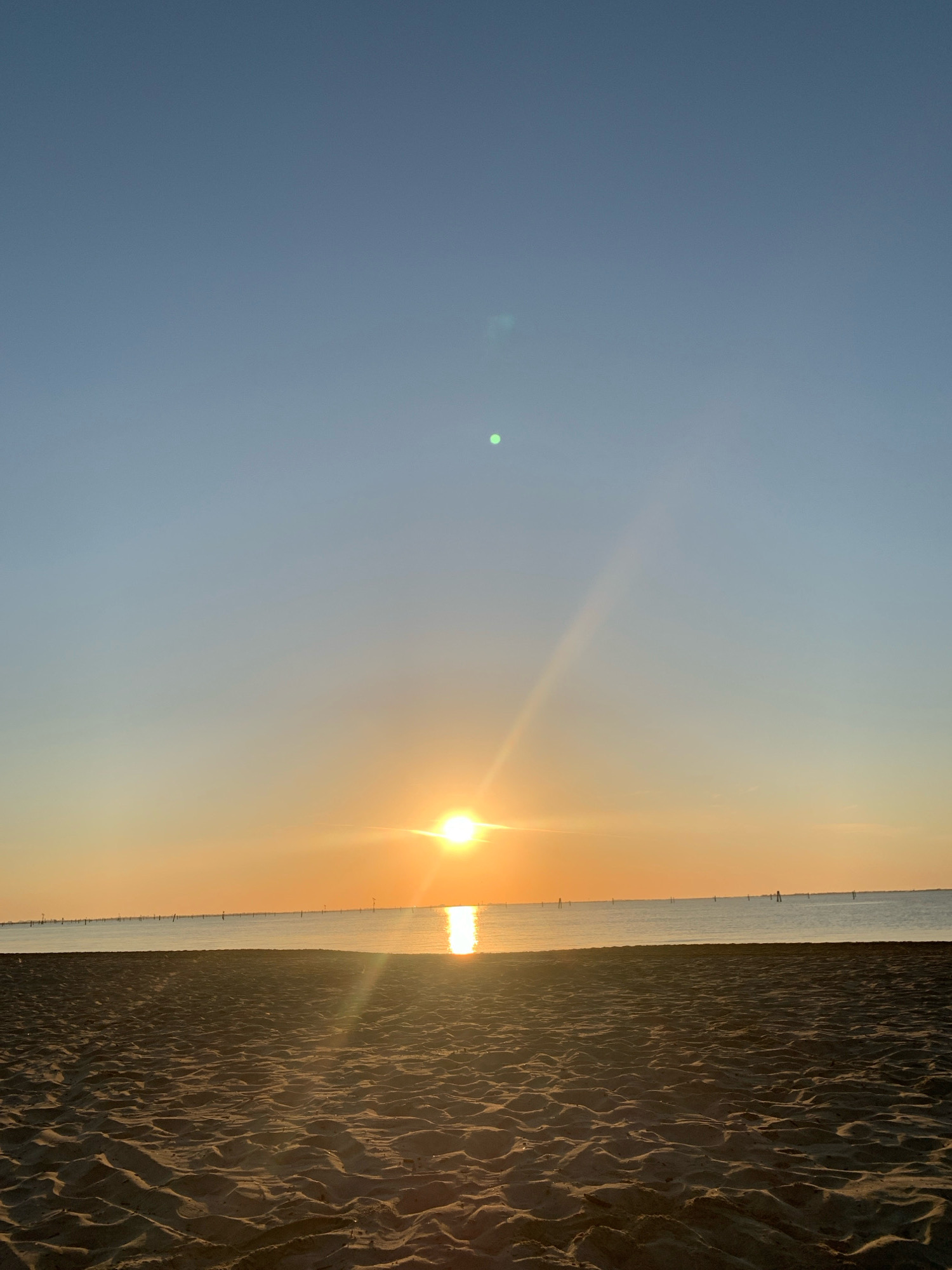 The image depicts a serene beach scene at sunrise or sunset. The foreground is dominated by textured sand, showing ripples and footprints. Beyond the sand, a calm body of water stretches to the horizon, reflecting the warm hues of the sky. The sun is positioned low in the sky, casting a bright glow and creating lens flare effects. The horizon is marked by a distant line of buoys or markers, and the sky transitions from a clear, light blue at the top to soft oranges and yellows near the horizon. The overall impression is one of tranquility and natural beauty. This scene is likely located on one of the beaches within the municipality of Comacchio, Italy, which is known for its extensive coastline and proximity to the Adriatic Sea.