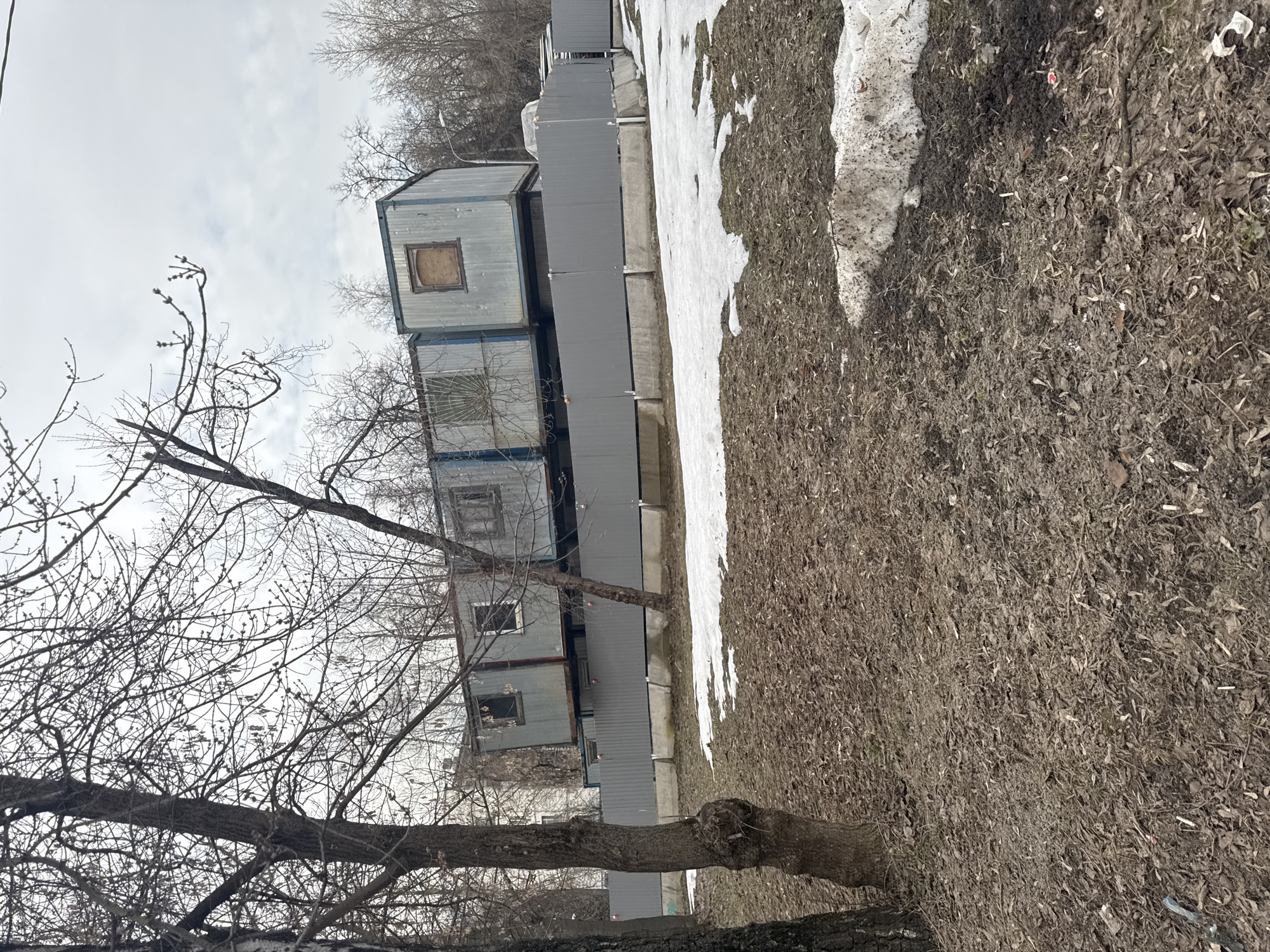 The scene depicts an outdoor urban environment in late winter or early spring at Aminevskoye Highway 12, Moscow, Russia, under an overcast sky.
In the foreground, the ground is composed of dark, damp soil intermingled with a dense covering of brown deciduous leaves and bare twigs. Several irregular patches of melting snow are present, particularly towards the right side. Two large, leafless deciduous trees with dark, textured bark are visible on the left, their branches extending upwards. No people or animals are present.
Across the midground, a gray, opaque panel fence, approximately 1.8 to 2 meters in height, extends horizontally. It is supported by concrete bases and features evenly spaced orange caps or markers atop its posts. Behind this fence, a complex of stacked, modular structures is visible. These structures, likely converted shipping containers or prefabricated units, are predominantly gray and light blue. They are arranged in at least two levels; some units exhibit a slight tilt or structural irregularity. Multiple units contain window openings: one is boarded with plywood, another features a distinctive decorative metal security grill, and others present empty frames or fragmented glass panes.
The background consists of dense, bare deciduous trees extending towards the horizon. A streetlamp with a white fixture is partially visible on the far right, above the fence line. No discernible actions or interactions are occurring within the visible area.