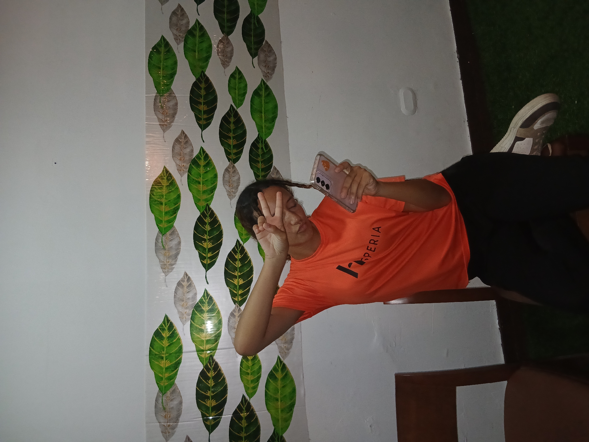 A young woman in an orange t-shirt and black pants poses for a selfie with her smartphone. She is making a peace sign with her left hand, held up near her face, eyes closed. The background behind her features a wall with a leaf pattern, painted in shades of green, yellow, and gray. To her right, there is a white wall. Her right shoe is visible, a brown and white sneaker. The scene is dimly lit, suggesting an indoor setting. The location is likely Playa El Agua, Venezuela, based on provided information, although the image itself does not contain explicit details that indicate it.