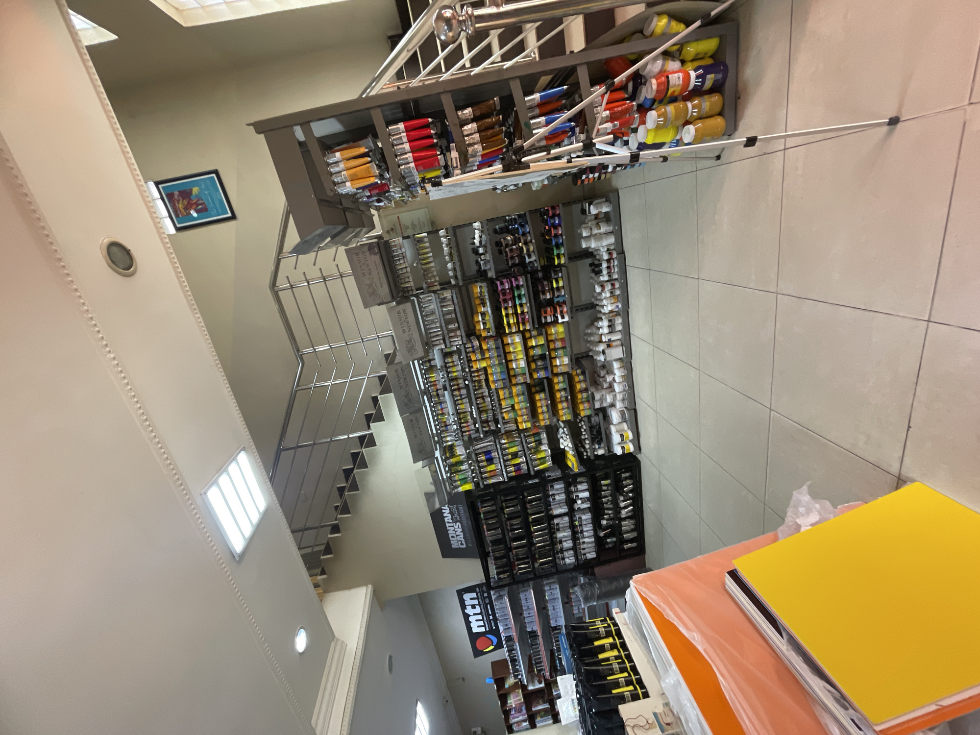 The image displays the well-lit interior of an art supply store, appearing clean and organized. On the right side, numerous shelves are densely packed with a wide array of art materials. Tubes of paint in various colors and sizes, likely oils or acrylics, are prominently displayed, some stacked horizontally, others in vertical slots. Product boxes bearing the names "WINSOR NEWTON" and "PLASTIM" are visible. Further down, large signs for "MONTANA CANS" and "mtn" indicate a section dedicated to spray paints.
On the left, a modern staircase with light-colored steps and sleek metal railings ascends, suggesting the building has multiple levels. A framed piece of abstract, colorful art hangs on the wall adjacent to the staircase. The walls are a light, neutral color, and the floor is composed of large, light-toned square tiles. The space is illuminated by several artificial light sources, including a rectangular fluorescent fixture on the ceiling and a circular light on the wall.
In the foreground, on the bottom right, there are stacks of large, flat sheets, likely art paper, cardstock, or foam boards, wrapped in plastic and presented in vibrant yellow and orange colors. Several long, white poles, possibly for painting or cleaning, are stored among the paint tubes on an upper shelf. No people are visible in the scene, which depicts a retail environment set up for customers. The store's clean and orderly presentation suggests it is well-maintained and ready for business.