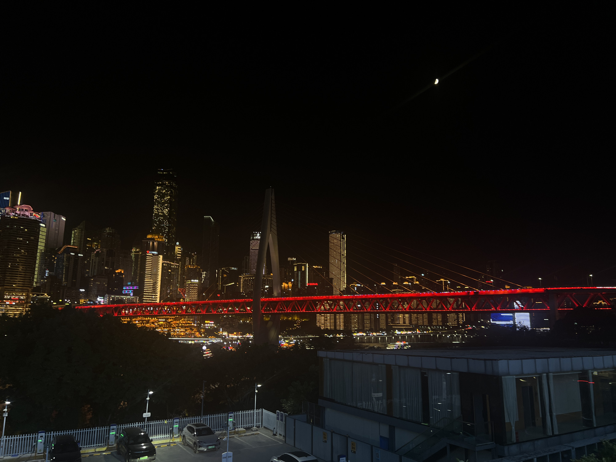 The image depicts a night view of a city skyline with a prominent illuminated bridge. The skyline consists of numerous tall buildings of varying heights and architectural styles, many of which are lit with colorful lights, creating a vibrant urban panorama. A large suspension bridge, painted red, spans across a body of water, with its illumination contrasting against the dark sky. The bridge is a central feature, drawing the eye with its glowing structure.
In the foreground, a modern building with large windows and a sleek design is visible, partially obscuring the view of the cityscape. In front of this building, a parking lot is set up with several charging stations for electric vehicles. Three electric cars are parked, two black and one silver, connected to the charging units. A white fence encloses the parking area. The environment appears to be a well-developed urban area, likely a commercial or residential district. The sky is dark, with a faint crescent moon visible in the upper right quadrant of the image, indicating it is nighttime. The city is located in Jiangbeicheng Subdistrict, Chongqing, China.