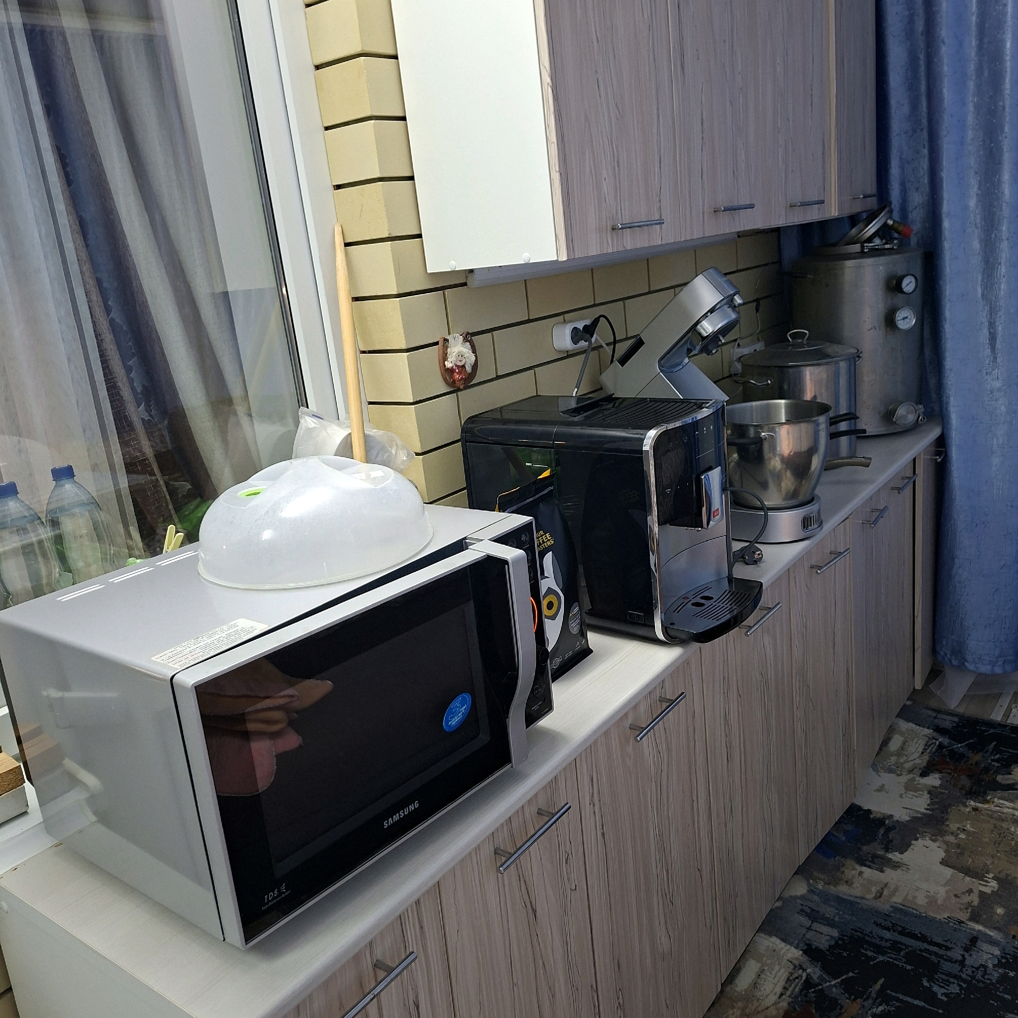 In a kitchen setting, a Samsung microwave is placed on a countertop. To its right, a black coffee machine sits next to a bag of coffee beans. Further to the right, a metallic pot rests on a silver heating element. Behind these appliances, two large metallic containers are visible, one with two pressure gauges and a red valve. The kitchen features light-colored tiled walls and wooden cabinets. A blue curtain hangs on the right side of the frame. On the left, through a window, two plastic bottles filled with water are partially visible. The floor is covered by an abstract patterned rug. The overall scene depicts a functional kitchen space. The location is Anapskaya, Russia.