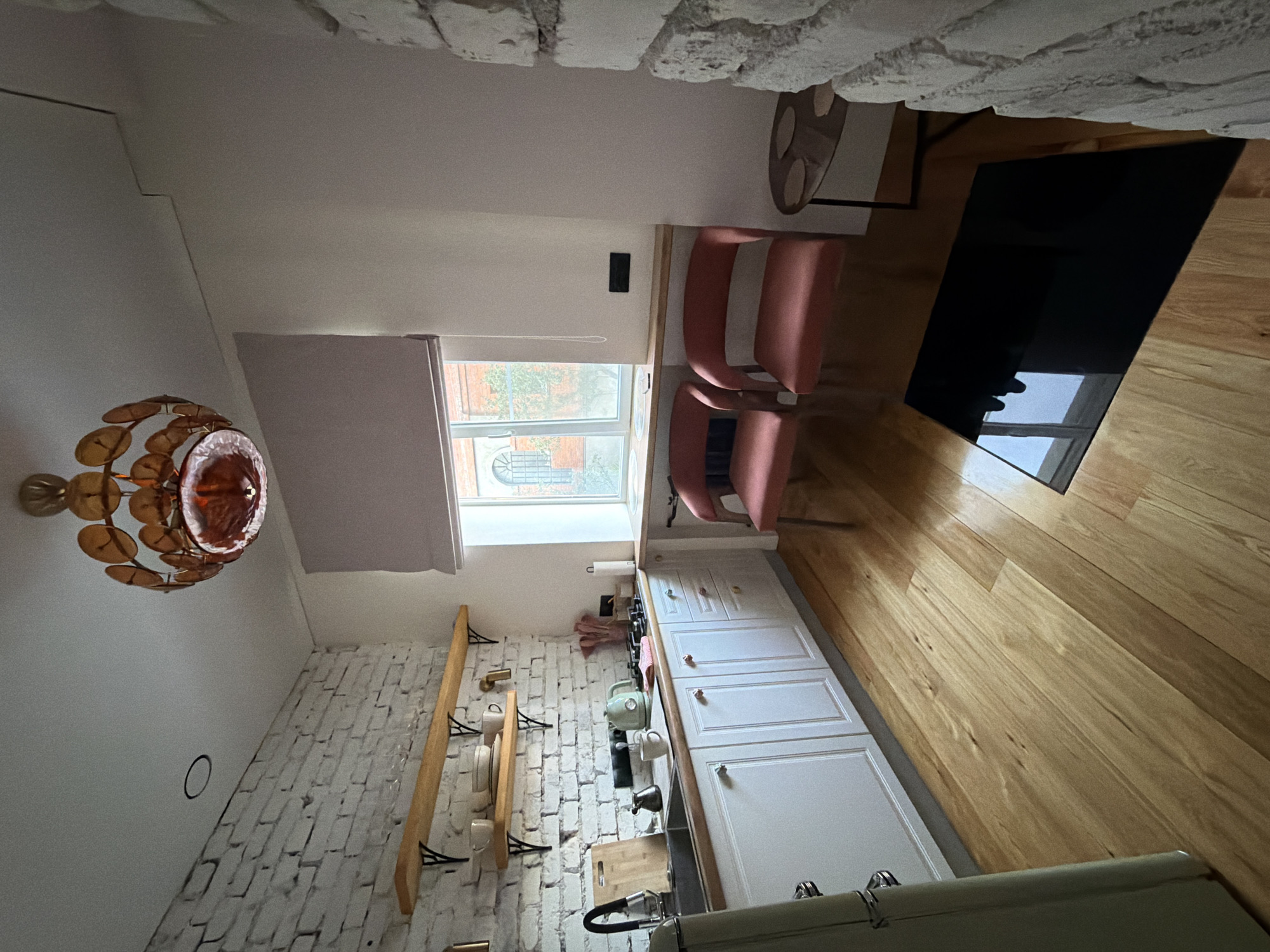 This is an interior shot of a compact, modern kitchen, possibly located in Kolomna, Russia, given the context. The kitchen features white brick walls on the left and right, contrasting with white cabinetry and a light-colored wooden floor. A wooden countertop runs along the length of the cabinets.
Above the sink area, there are two light-colored wooden shelves with black metal brackets, displaying a few white ceramic dishes and cups. The sink itself is stainless steel and has a black faucet. To the right of the sink, on the countertop, is a green electric kettle, a small black coffee pot, and a mug.
A large window with a light purple roman blind dominates the back wall. Below the window, there are two coral-pink upholstered chairs. A small round side table with a dark top and thin legs is positioned to the right of the chairs. In the foreground, a black rectangular object, likely a stovetop or a mat, is placed on the wooden floor.
A distinctive chandelier with a circular frame and numerous wooden disc-like elements hangs from the white ceiling. The overall impression is a small, functional, and stylish kitchen space.
