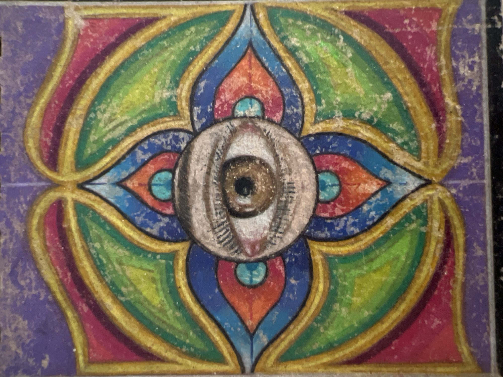 A rectangular image features a stylized eye at the center of a symmetrical, lotus-like design. The design consists of concentric layers of teardrop shapes in purple, pink, green, blue, and orange, all outlined in gold. The eye is depicted in shades of brown and white. The image has a worn or aged appearance. The location in the city of Madrid, Spain is not present.