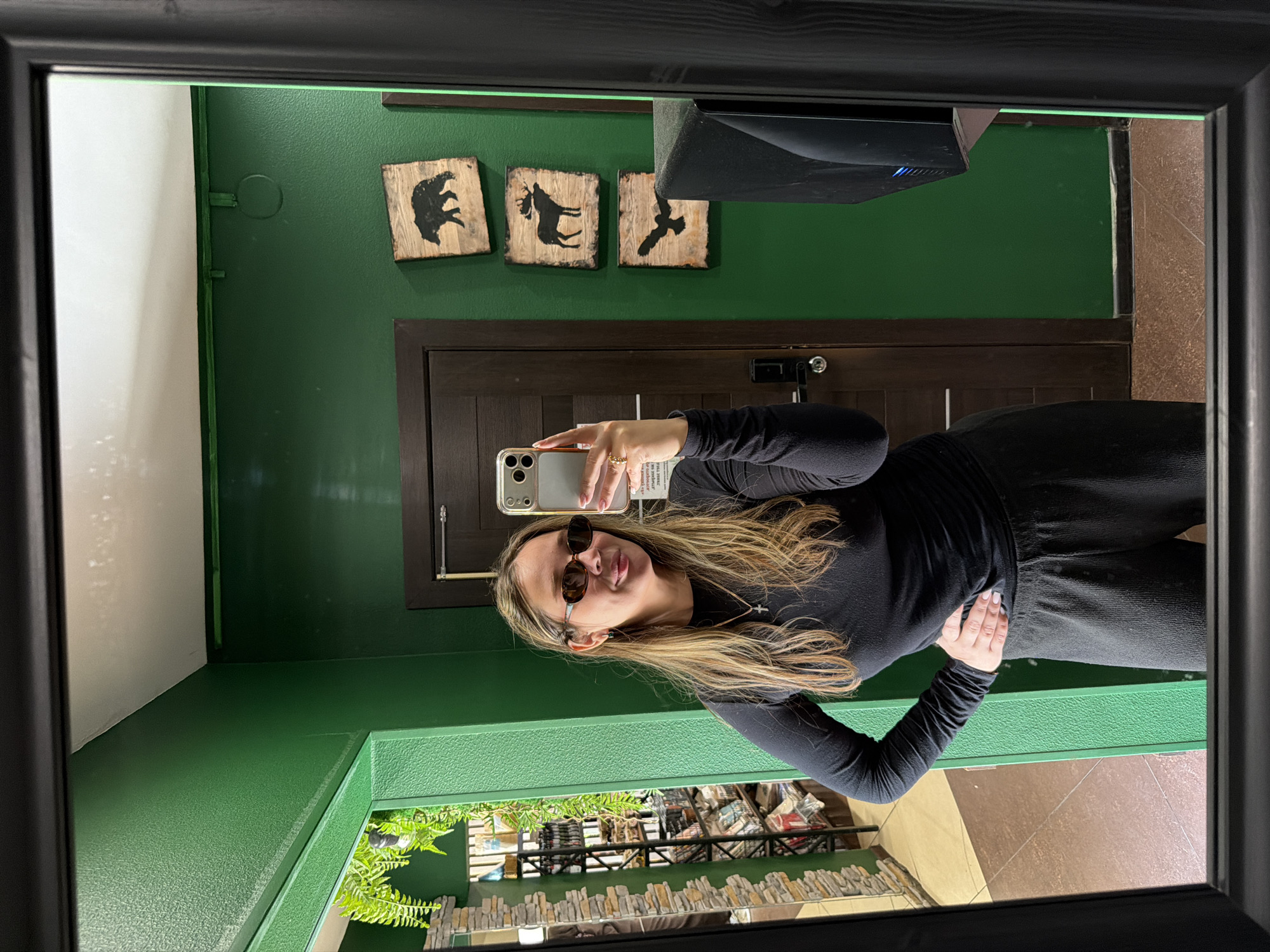 A woman is taking a mirror selfie in a room with green walls. She has long, blonde hair and is wearing sunglasses, a black long-sleeved shirt, and black sweatpants. She is standing with one hand on her hip. In the background, there is a dark brown door and three wooden plaques with silhouette images of a bear, a moose, and a bird. To the left of the woman, a shelf unit with various items is visible in the reflection of another mirror. The overall scene appears to be an indoor space, possibly a shop or a home, located in the city of Sortavala, Russia.