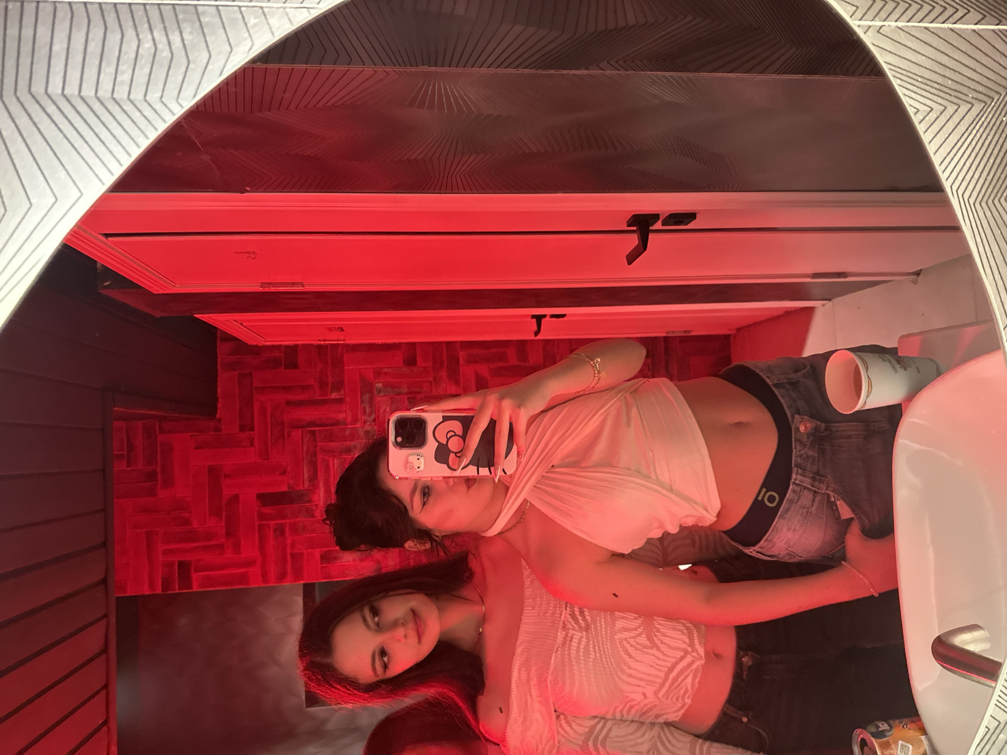 The image displays two young adult females inside a public restroom, likely within a commercial establishment in Yerevan, Armenia. The scene is illuminated by strong red ambient lighting.
In the foreground, a white ceramic sink basin with a chrome faucet is visible. Resting on the sink's edge are a silver-colored beverage can with an orange and white label, and a light-colored mug.
The primary subject, on the right, has dark hair secured upwards and wears a white halter-neck crop top that exposes her midriff, paired with blue jeans. She holds a light-colored smartphone, adorned with a "Hello Kitty" case, directed towards the mirror to capture a selfie. A gold-colored bracelet is present on her left wrist.
The secondary subject, positioned behind and to the left of the primary subject, has long, dark hair and wears an off-the-shoulder white top with a textured pattern, along with dark-colored jeans. She is observed looking to her right. A thin gold-colored necklace is visible around her neck.
The background wall behind the subjects features a red, textured surface with a horizontal brick or tile pattern. To the right, three white doors with black handles are visible, also bathed in red light. The right edge of the mirror shows a grey and white zigzag pattern. The ceiling appears dark.