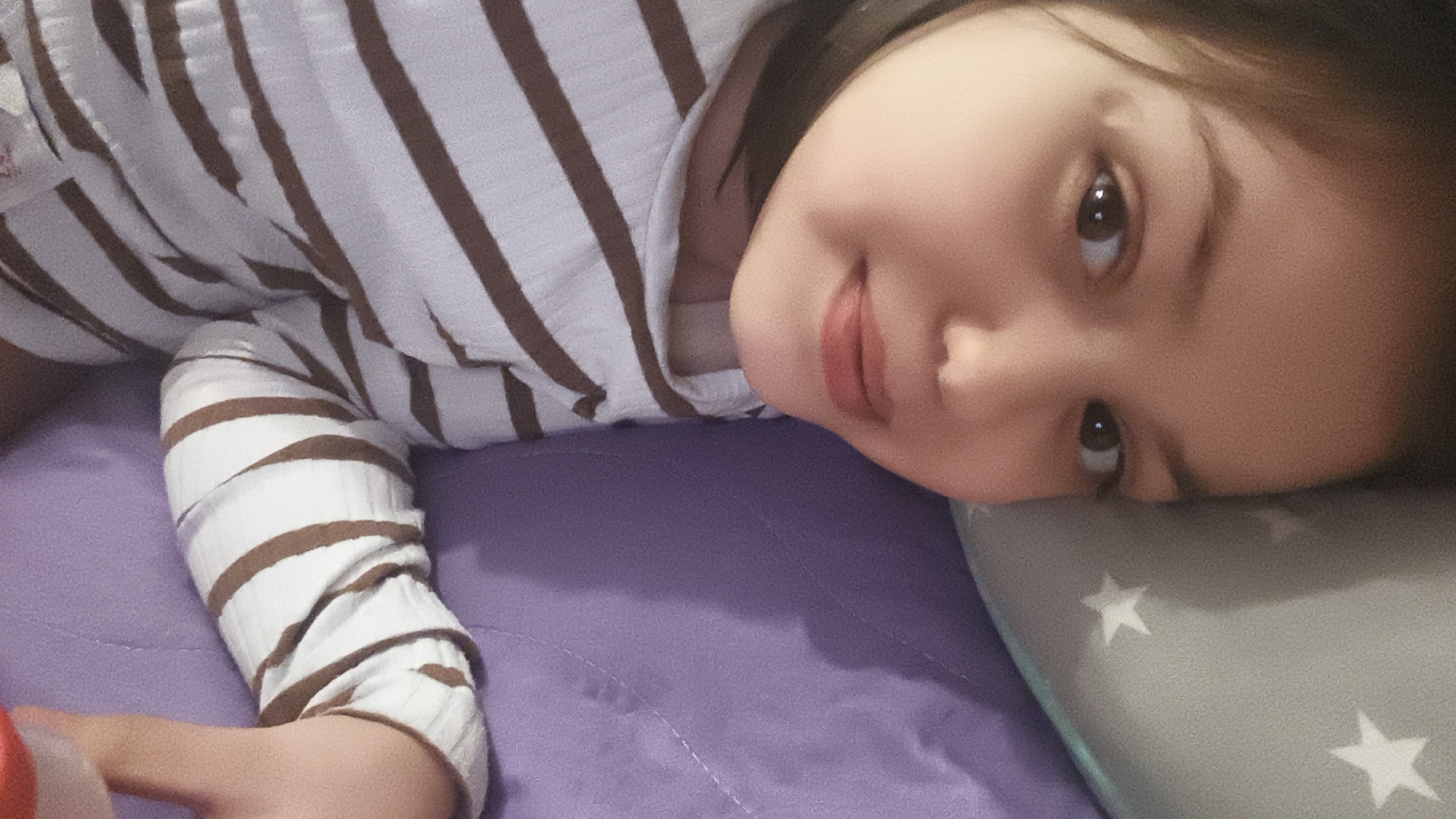 A child is lying on their side on a purple bed and a gray pillow with white stars. The child has brown hair and brown eyes, and is wearing a white and brown striped shirt. Their arm is extended in front of them.