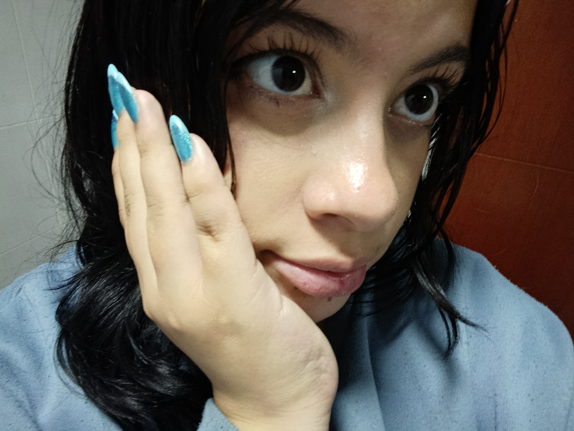 A close-up, indoor shot of a young person, likely female, looking directly at the camera with wide, dark eyes. Her dark hair, which appears wet or styled to look wet, frames her face and shoulders. She wears a light blue or greyish garment, possibly a sweater or hoodie. Her left hand, adorned with long, pointed, glittery light blue nails, rests against her cheek and jawline, partially obscuring her face.
Her expression suggests intense focus, curiosity, or slight surprise. Her skin exhibits a natural sheen, particularly on her nose and forehead, and her lips are slightly parted. The background consists of a light-colored wall on the left and a reddish-brown wall on the right, indicating an indoor setting. There is no visible text in the image. The image is set in Funza, Colombia.