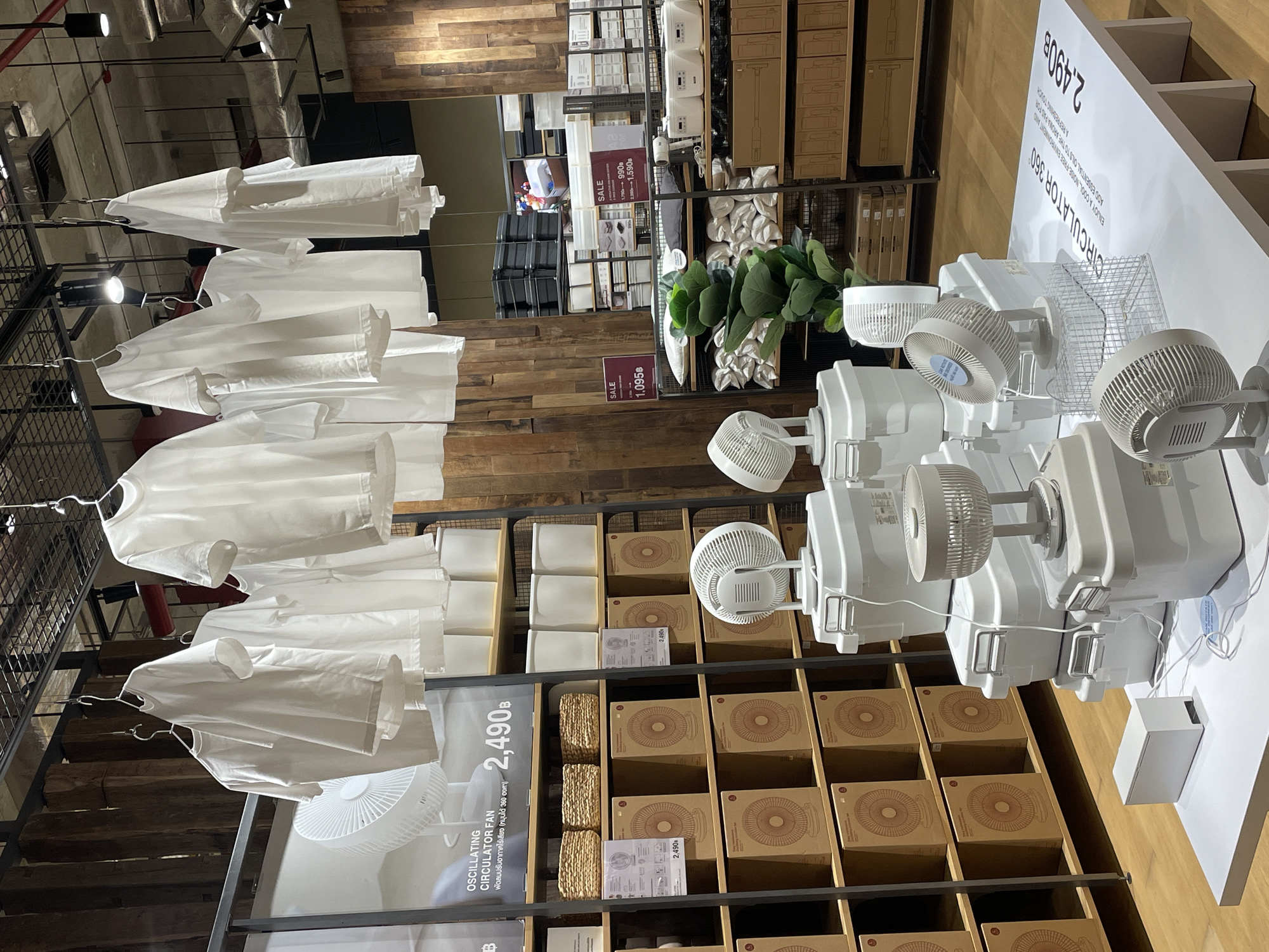 This is an interior shot of what appears to be a MUJI store in Bangkok, Thailand. Rows of white shirts hang on display racks. In the foreground, oscillating circulator fans are exhibited on a shelf. Signage near the fans indicates a price of 2,490 Baht. The shelves underneath the fans hold the fans’ packaging. Behind the shirts and fans, there is a wooden wall and shelving displaying other MUJI products. There are no people visible in the image.