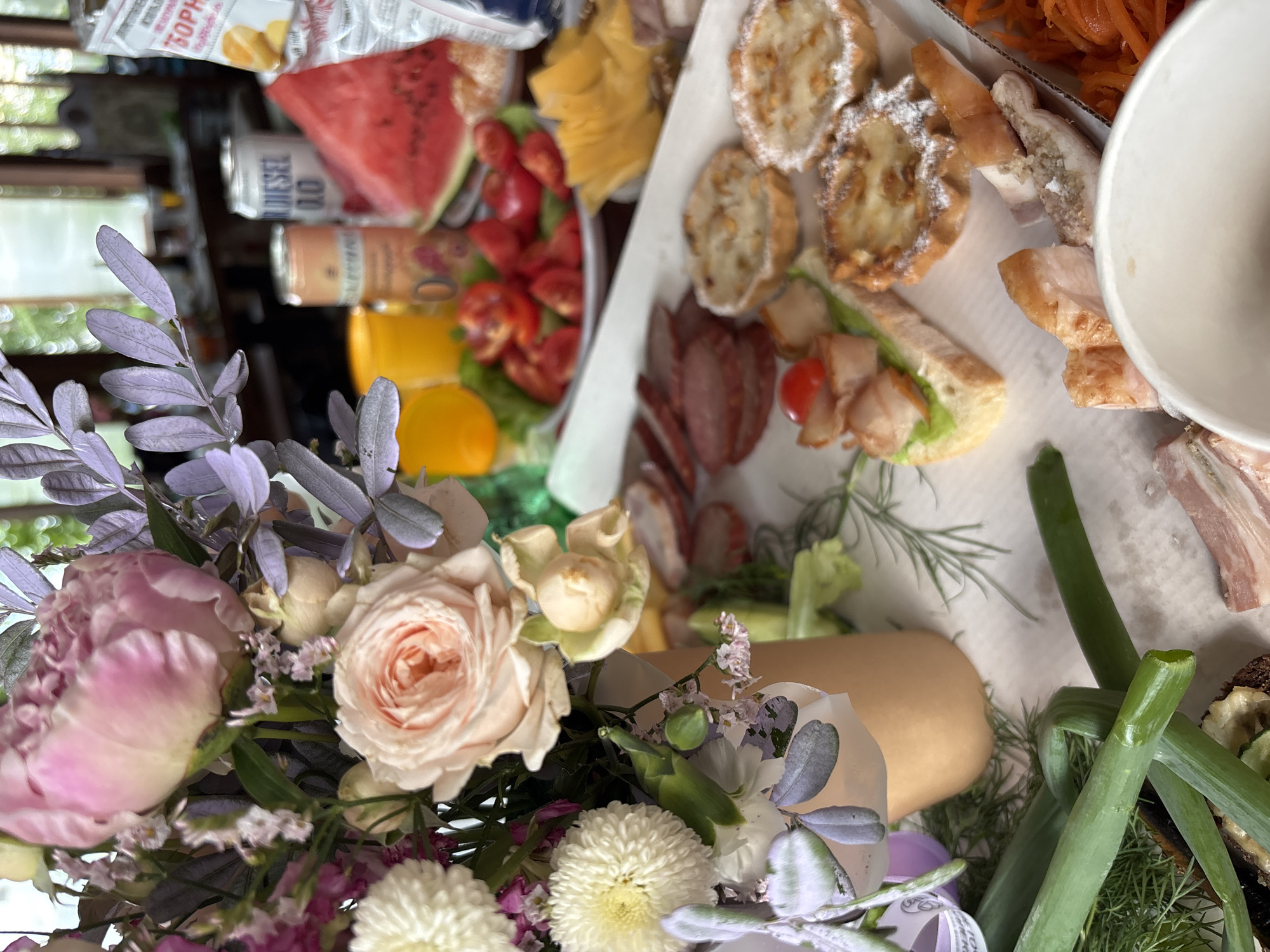 A close-up, overhead shot captures a table laden with food and drinks, set outdoors with a hint of a building in the background. The scene is brightly lit, suggesting daytime. In the foreground, a bouquet of flowers with pink and peach roses, white chrysanthemums, and purple foliage is arranged in a peach-colored vase. To the right of the flowers is a platter of assorted foods, including sliced meats, small savory tarts dusted with powdered sugar, and small open-faced sandwiches topped with cherry tomatoes and lettuce. Further back, a watermelon slice, tomatoes and lettuce in a bowl, sliced pineapple, and two beverage cans are visible. The overall impression is of a celebratory gathering or picnic. The provided location of Chaikovsky, Russia, is not visually verifiable within the image itself.