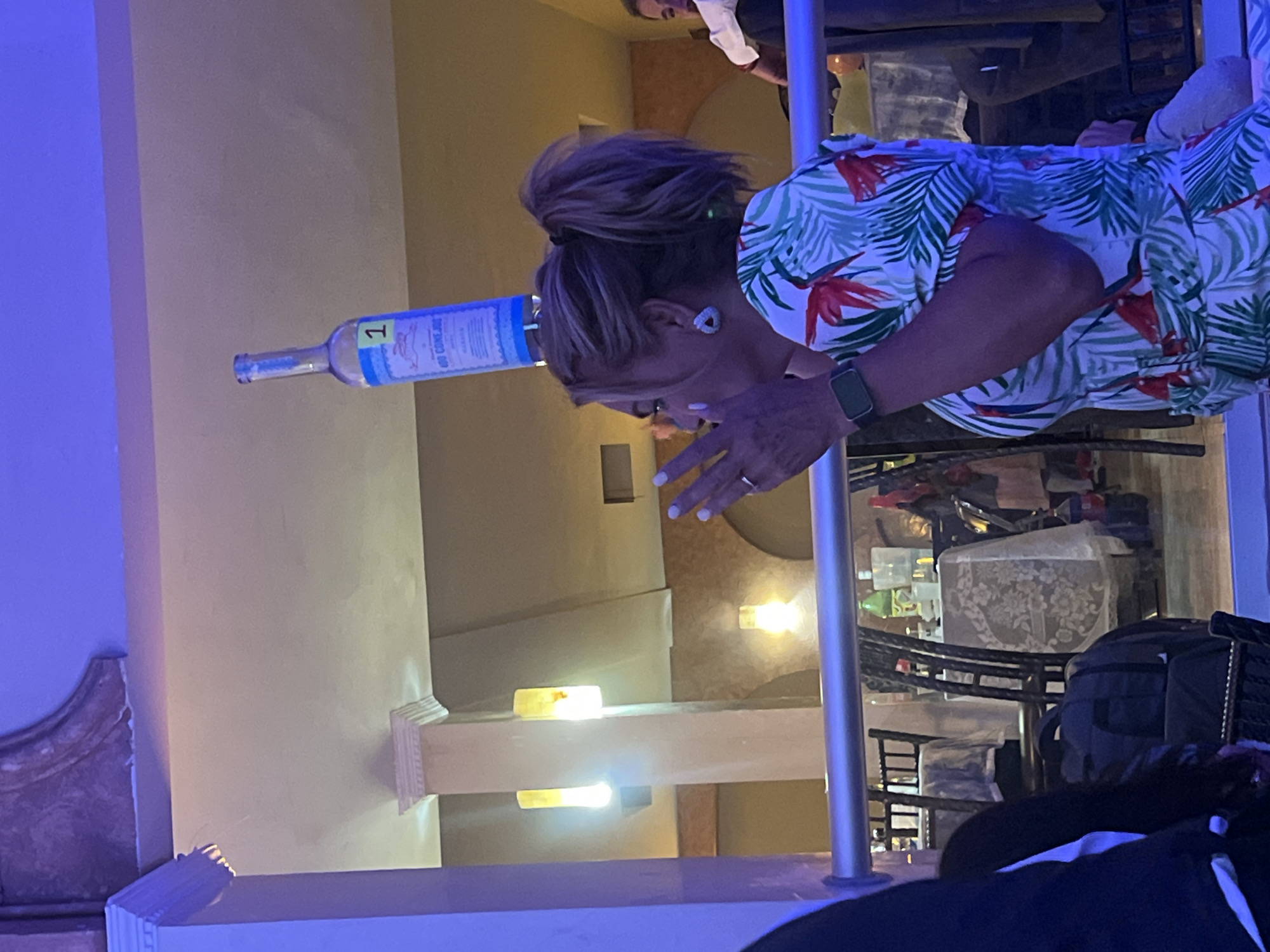A woman with brown hair in a high ponytail is attempting to balance a blue wine bottle on her forehead. She wears a white shirt with a tropical print. Her left arm is visible, adorned with a smartwatch. She is at a gathering in the city of Puebla, Mexico. A chrome pole is near her. Another person in a white shirt is in the background. Bags are visible in the background.