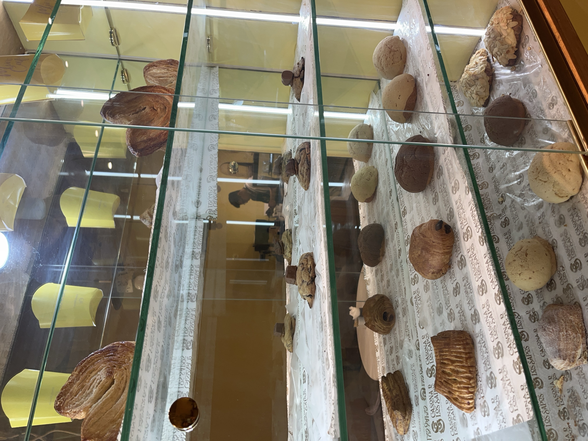 The image depicts a bakery display case with various pastries. The pastries include palmiers, cookies with chocolate pieces, and round, crusty rolls. The background appears to show the interior of a store, with a reflection of people visible in the glass. The pastries are arranged on printed paper with the words "Casona de Los Sapos", suggesting the location is likely in Puebla, Mexico, a city known for its landmark, Casona de Los Sapos. The overall impression is that of a bakery or pastry shop selling a variety of baked goods.