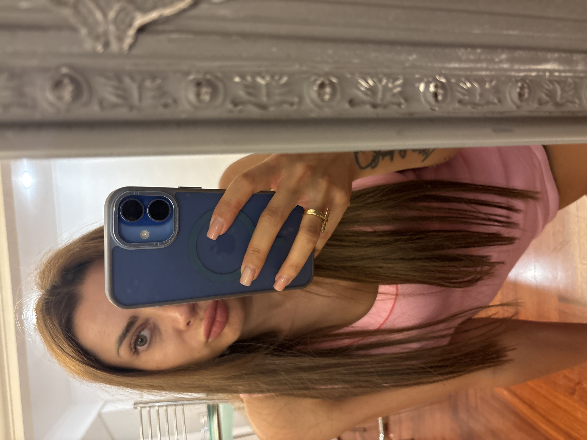 A woman with long, brown hair and light skin is taking a selfie in a mirror. She is wearing a pink tank top and has a gold ring on her left hand. She is also wearing false eyelashes and has a tattoo on her left arm. The mirror appears to be ornate and is reflecting part of a room with wooden floors and a silver towel rack. There is a smartphone being held in her right hand, obscuring part of her face. The background is blurred, but it appears to be an indoor setting, possibly a bathroom or dressing room. The scene is captured from the chest up. There is no indication of the location being Rome, Italy.