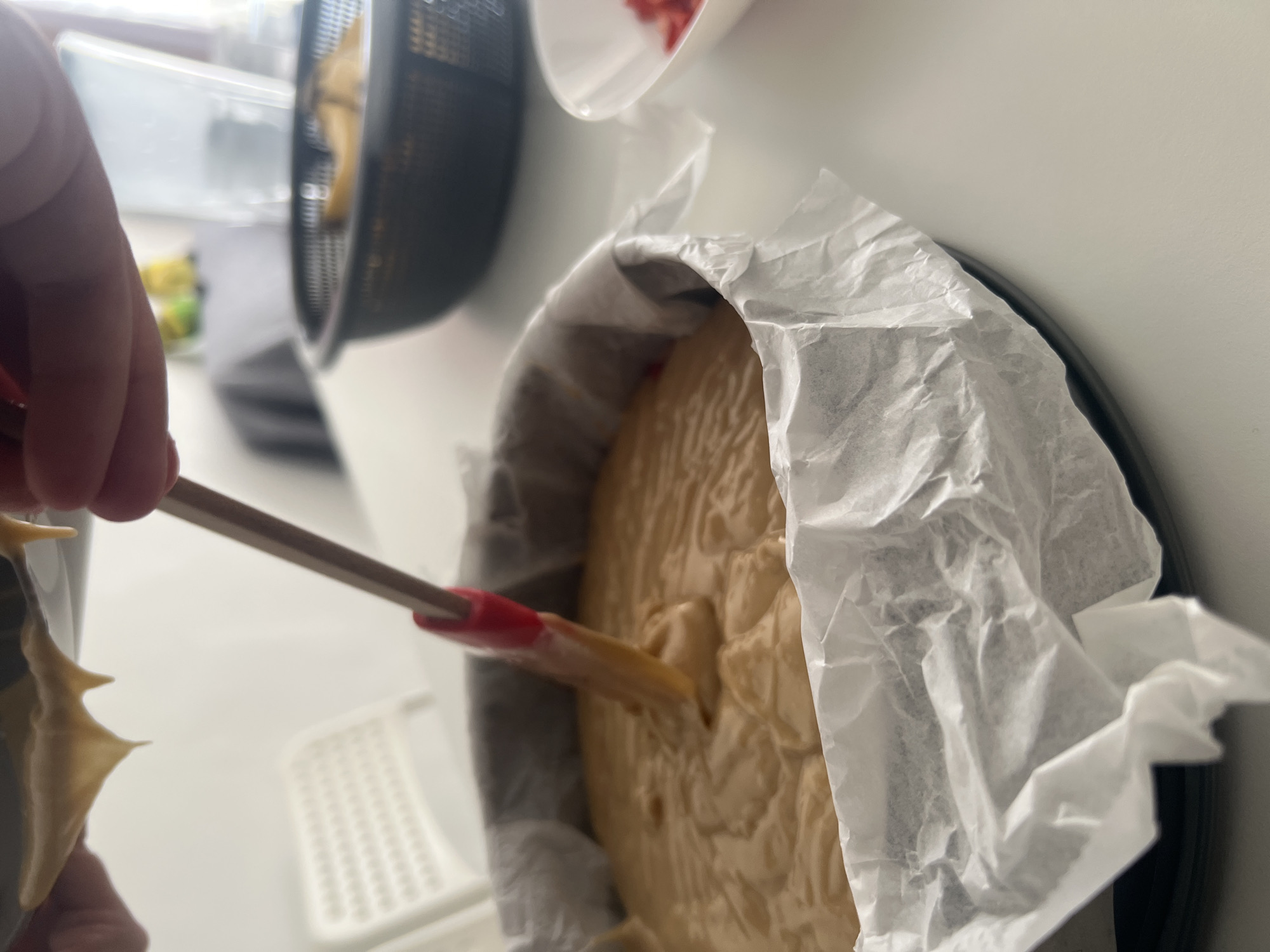 The image shows a person preparing a cake in a kitchen. The cake batter is in a round cake pan lined with parchment paper. The person is using a spatula to spread the batter evenly. The background shows other kitchen items. It is impossible to determine the location in Mogán, Spain from the image.