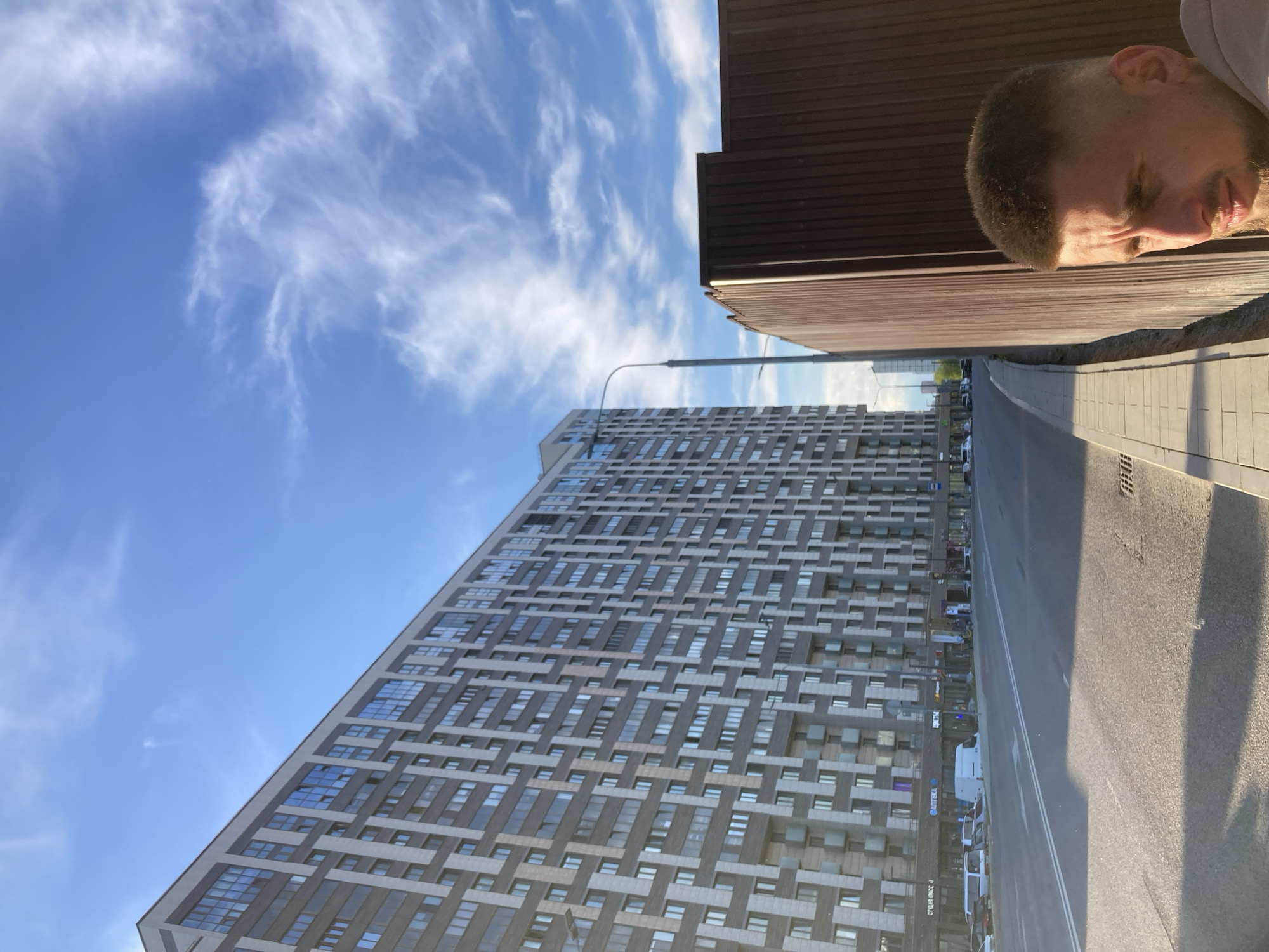 This image captures an urban street scene in Moscow, Russia, bathed in the light of either an early morning or late afternoon sun, as evidenced by the long shadows cast across the pavement. The sky is a vibrant blue, adorned with delicate, wispy white clouds, indicating clear weather.
The left side of the frame is dominated by a towering, modern building with a distinctive geometric facade in shades of grey and brown, featuring numerous windows. Below it, a paved road stretches, marked by a double white line, with several cars parked along its side. A street light pole extends upwards from the pavement near the road. On the right, a patterned sidewalk leads to a partially visible brown building, possibly featuring corrugated metal siding.
In the upper right corner, a man's head and shoulder are visible, as if he is looking down onto the scene. He has short, dark hair and stubble, and his expression appears calm or contemplative, with his eyes seemingly closed or cast downwards. He wears a light-colored garment.
Visible text on signs at the base of the tall building includes "АПТЕКА" (Pharmacy) and a partially obscured sign reading "СУШИ КРОСС" (Sushi Kross), providing specific commercial details of the location. The overall setting depicts a contemporary city environment.