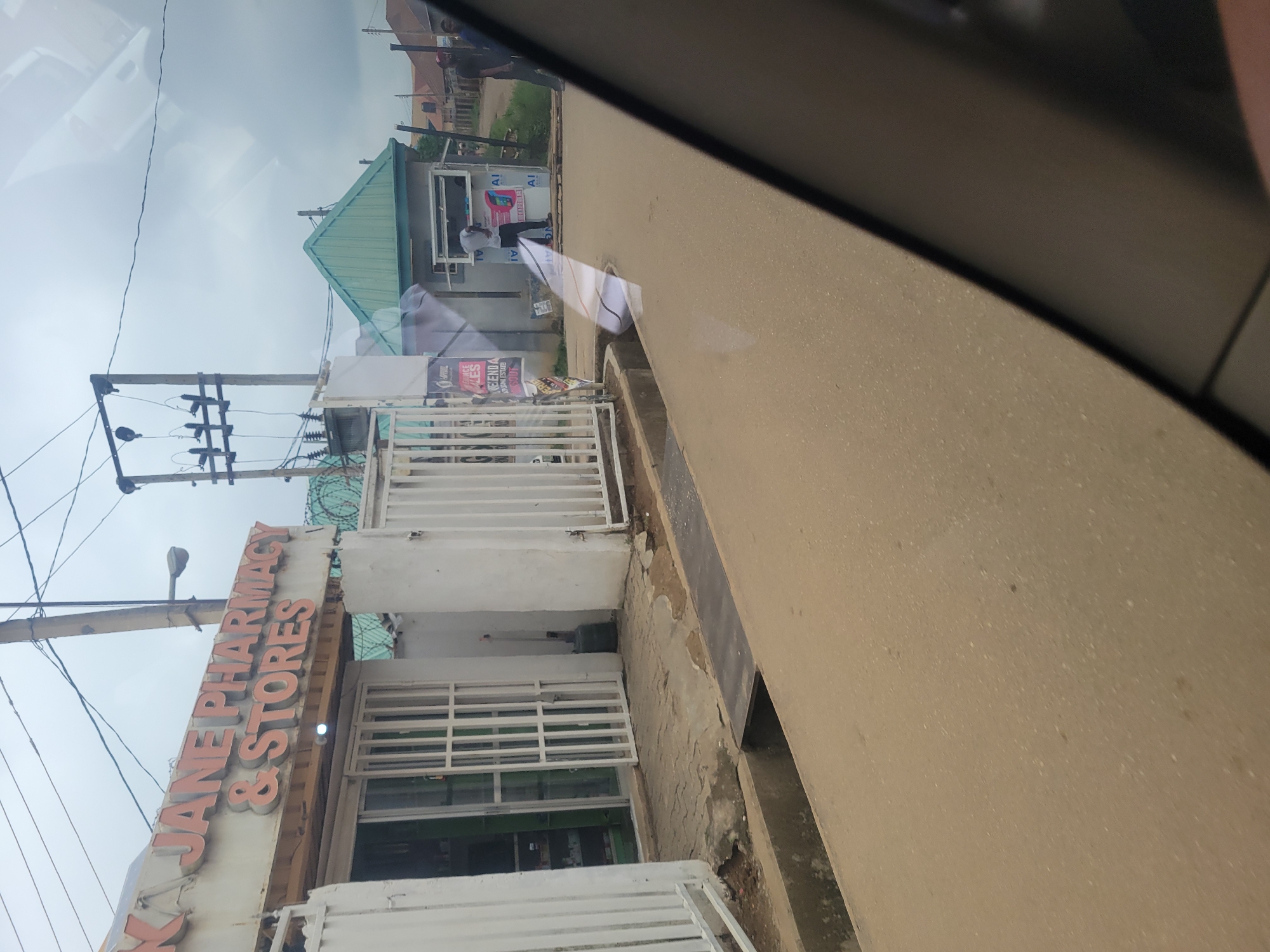 The image depicts a street scene in Katambe, Nigeria. In the foreground, a tan-colored road with scattered debris occupies the lower half of the frame. A concrete drainage channel runs along the edge of the road. To the left, a white building with metal security gates is visible. Above the entrance, a sign reads "AJANE PHARMACY & STORES" in orange lettering. Inside the pharmacy, shelves stocked with various items can be seen through the glass. Adjacent to the pharmacy, a similar white building with white metal gates extends further. Further down the street, to the right, a smaller green-roofed structure serves as a kiosk or small shop. A person in a white shirt is visible standing in front of this kiosk, seemingly interacting with it. Other people are present in the background, some walking and others standing near buildings. Electrical poles with wires are prominent in the sky. The overall environment appears to be a commercial area with a mix of residential and business establishments. The sky is overcast.