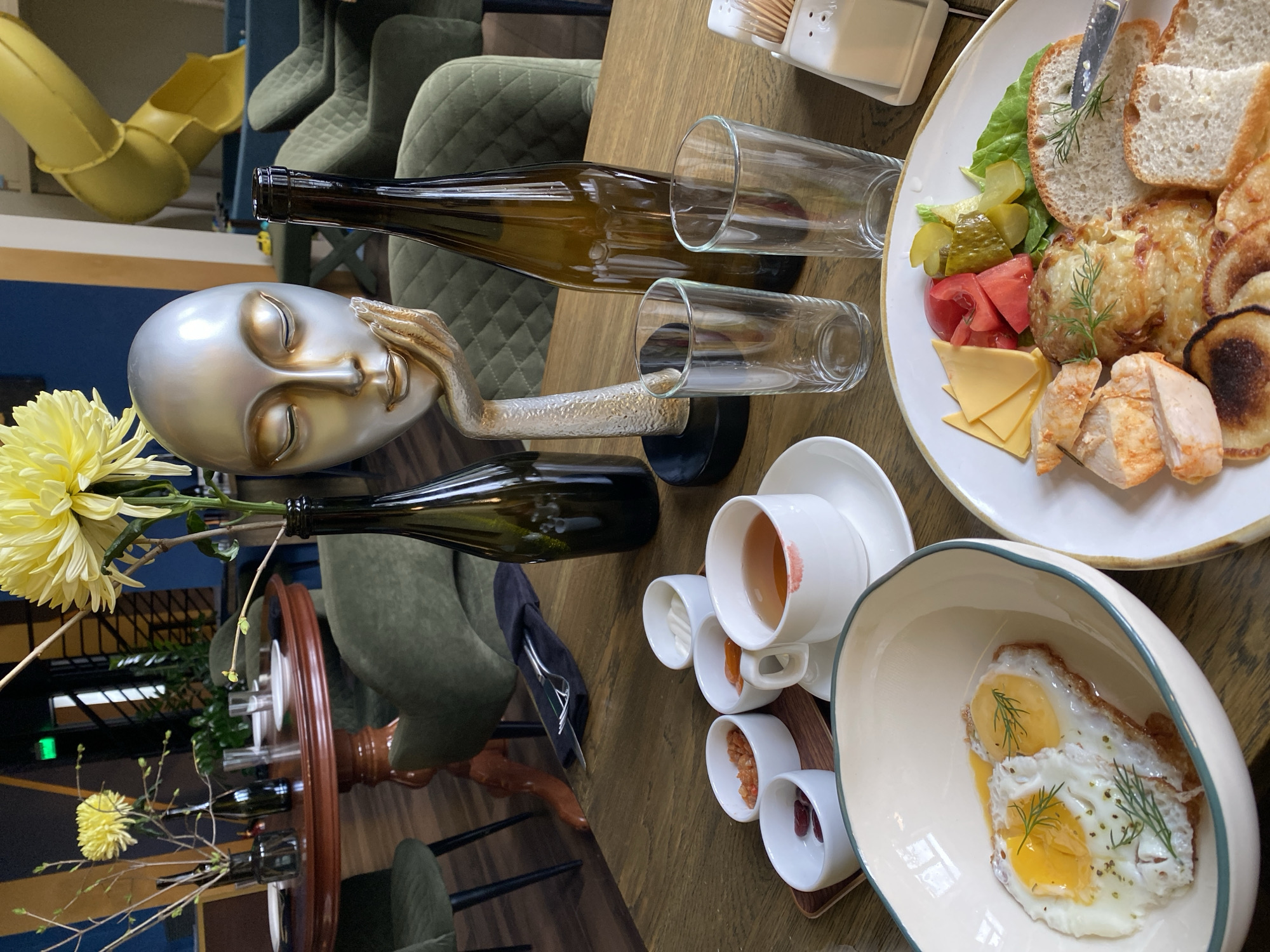 A table in a restaurant in Yekaterinburg, Russia, is set with various items. There are two wine bottles lying on their sides next to two drinking glasses. There is a sculpture of a silver head with a hand covering its face. There is a cup of tea, with a lipstick stain on the rim, surrounded by small bowls of condiments on a wooden tray. A plate with sliced bread, cheese, pickles, tomatoes, and fried food is present. A bowl contains two fried eggs with dill. There is a bouquet of yellow chrysanthemums. The table is wooden and surrounded by green upholstered chairs.