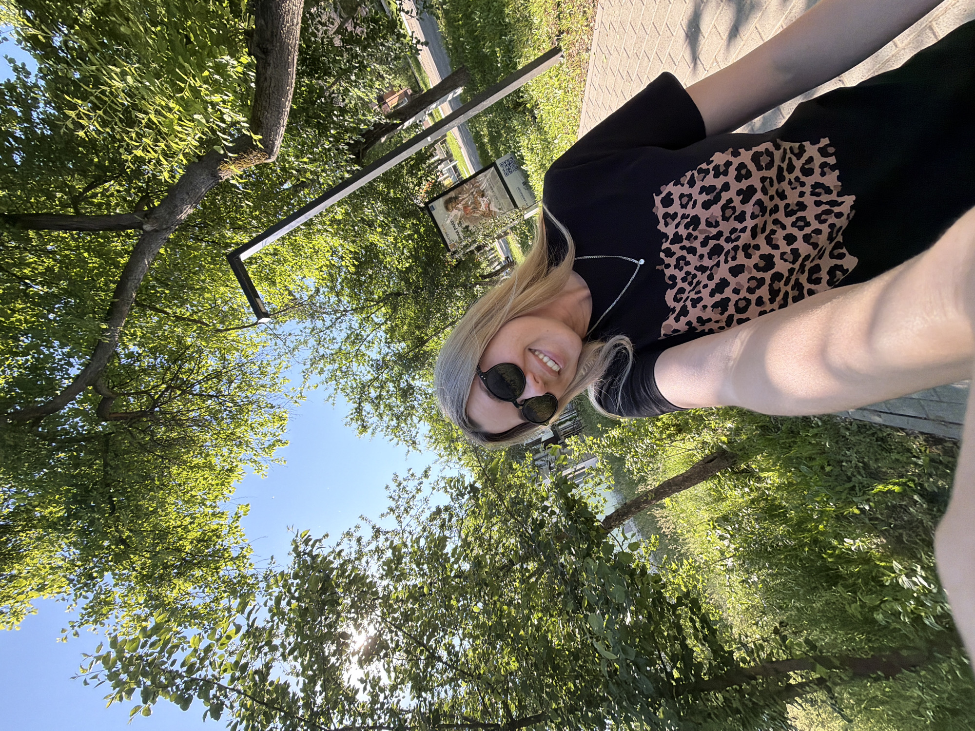 In Chaikovsky, Russia, a woman with blonde hair and sunglasses takes a selfie outdoors on a sunny day. She is wearing a black t-shirt with a leopard print design and a silver necklace. The background features lush green trees and foliage, a paved walkway, and a lamppost. A poster is visible on a stand in the background. The sun is shining brightly, creating dappled light through the leaves.