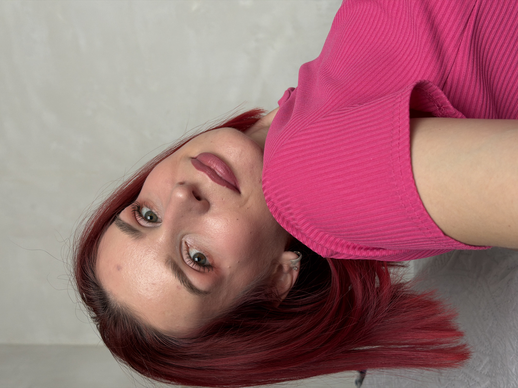 A woman with red hair and green eyes, wearing a pink ribbed top, is shown in a close-up shot. She is looking directly at the camera. A silver earring is visible in her left ear. The background is a plain, light gray wall. There is no indication that this image was taken in the city of Chaikovsky, Russia, nor are there details suggestive of any particular location.