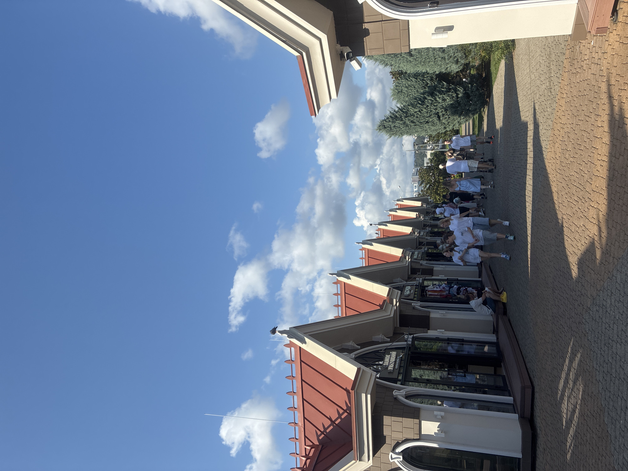 A street scene in Sochi, Russia, depicts a row of red-roofed market stalls with brown and cream facades. The stalls are lined up along a cobblestone pathway, with a few people walking and browsing. One man sits on a ramp in front of a stall. Green cypress trees line the pathway on the right side of the image. The sky is blue with scattered clouds, and the sun casts shadows on the ground.