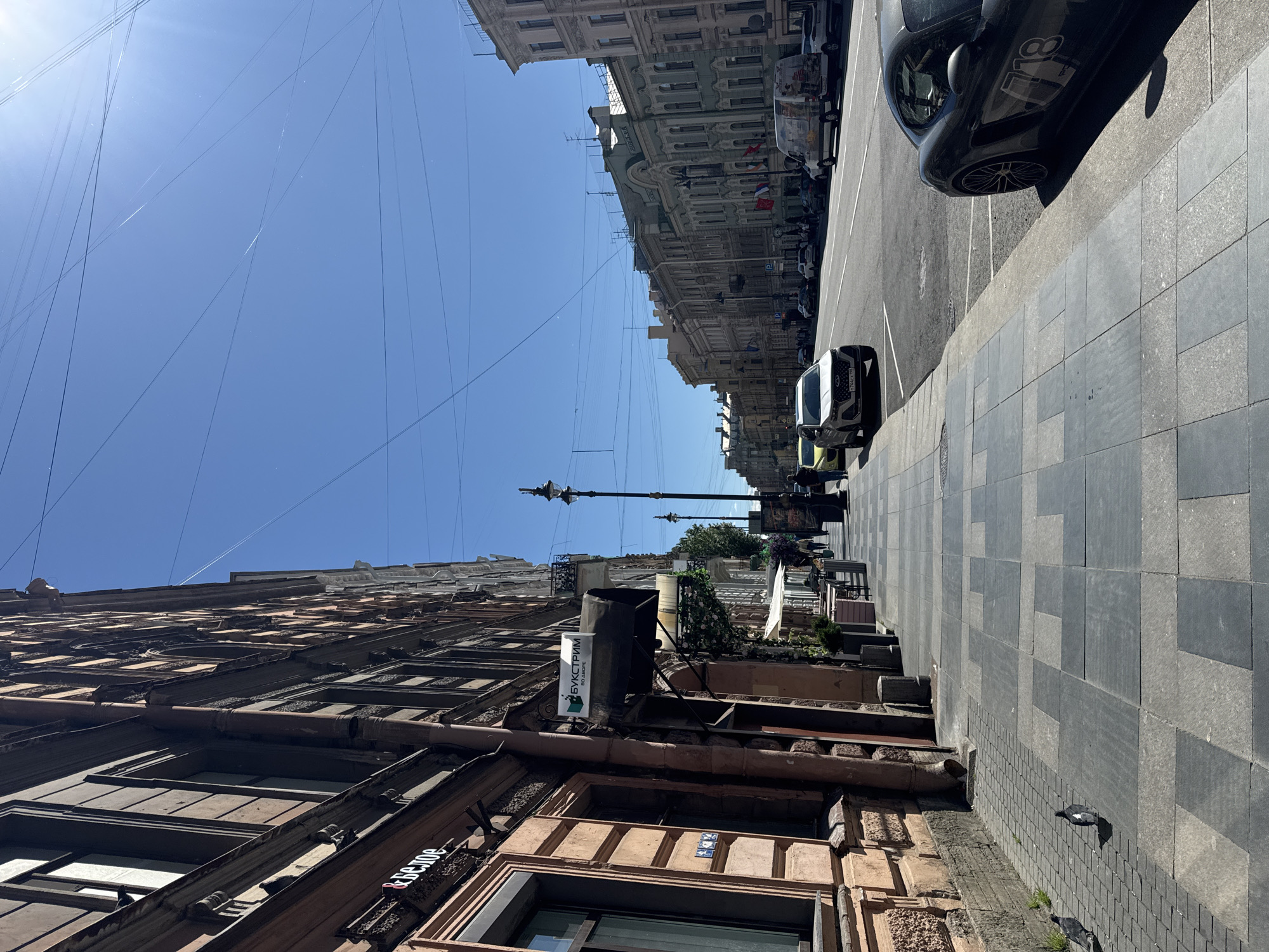This image depicts a street scene in Saint Petersburg, Russia, on a clear, sunny day. The perspective is from a low angle, looking upwards and along the street. On the left, aged buildings with ornate facades and numerous windows line the street, their architectural details suggesting a historical European city. Wires crisscross the bright blue sky above.
On the right side of the street, a paved sidewalk runs alongside parked cars. One dark car with the number "78" on its side is prominently featured. Further down the street, other vehicles and some pedestrians are visible. The buildings on this side appear grand and well-maintained, with flags displayed from some of them. The sidewalk is paved with large, grey tiles arranged in a geometric pattern. A pigeon is seen on the pavement near a wire fence.
A sign with Cyrillic text that appears to read "БУКСТРИМ" is attached to one of the buildings. The overall atmosphere is bright and daytime, with strong sunlight casting shadows, indicating a time around midday. The scene conveys a sense of urban life in a historic city.