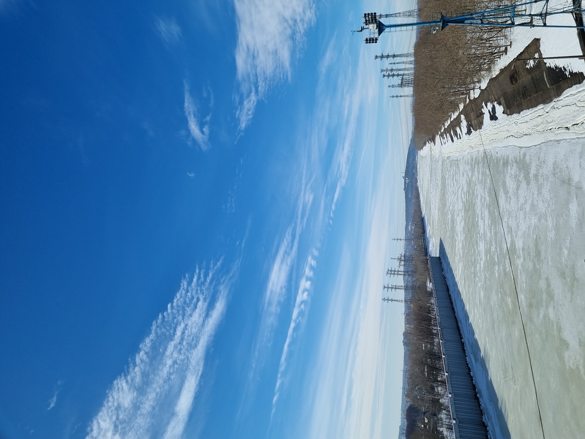 This image depicts a wide, frozen waterway, likely a canal, in the city of Chaikovsky, Russia, observed from an elevated perspective on a clear winter day. The sky is deep blue with scattered, elongated cirrus clouds. The waterway itself is entirely covered in a thick layer of patchy ice and snow, extending into the distant horizon.
On the left side of the waterway, a long, vertically corrugated metal retaining wall runs parallel to the ice. Beyond this wall, a line of deciduous trees, devoid of leaves, precedes a distant urban landscape featuring various buildings. Several large lattice-style electricity transmission towers are visible on the far-left horizon.
The right bank of the waterway features a sloping, snow-dusted brown earth embankment. A dense line of bare deciduous trees runs along this bank, also extending into the distance. Multiple large lattice-style electricity transmission towers are prominently positioned along this right bank in the mid-ground and background. In the immediate foreground on the right, a tall, blue metal tower, equipped with multiple floodlights or instrumentation near its top, stands partially within the frame. A dark cable or line stretches horizontally across the frozen waterway from the vicinity of this blue tower towards the left bank. There are no individuals, vehicles, or apparent activities present in the scene.