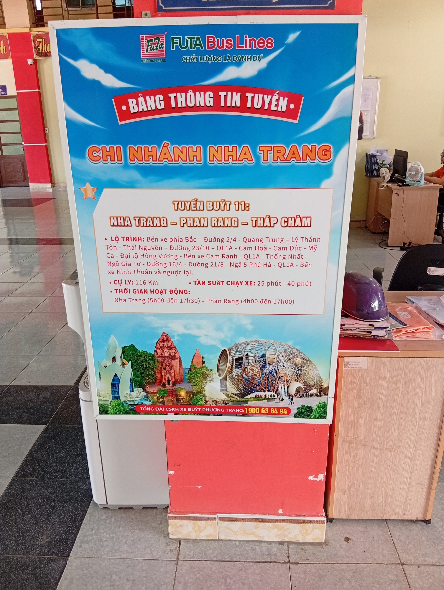 The image shows a sign for FUTA Bus Lines, specifically a route information board for the Nha Trang branch in Phan Rang – Tháp Chàm, Vietnam. The sign is predominantly blue and white, featuring the FUTA Bus Lines logo at the top. Below the logo is the text "BẢNG THÔNG TIN TUYẾN" ("Route Information Board") and "CHI NHÁNH NHA TRANG" ("Nha Trang Branch"). The sign details Route 11: Nha Trang - Phan Rang - Tháp Chàm, including the route itinerary, distance (116 Km), and the frequency of bus service (25-40 minutes). Operating hours are listed as Nha Trang (5:00 to 17:30) and Phan Rang (4:00 to 17:00). The bottom of the sign includes a contact number for customer service: 1900 63 84 94. Below the text are images of landmarks in the area. The sign is positioned in front of a reception desk and other pieces of furniture inside an office area.