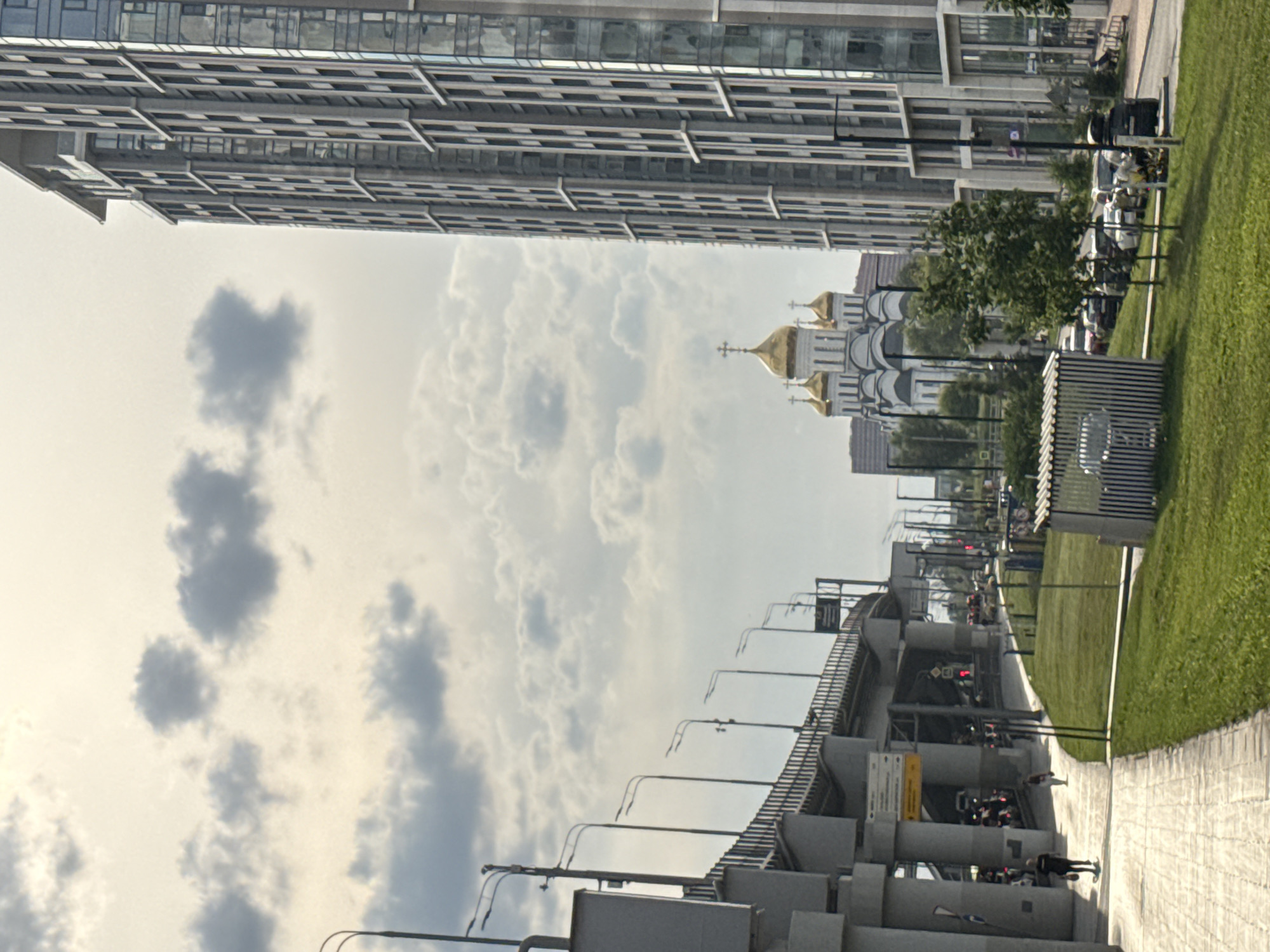 The image depicts a street scene in Moscow, Russia, on a cloudy day. On the right is a tall, modern high-rise building with a glass facade and numerous windows. To the left of the building and further in the background, a white church with golden domes is visible. In the foreground on the left, a multi-level overpass structure with concrete pillars and railings is prominent, with several streetlights lining it. A paved pedestrian walkway runs alongside a grassy area. Several people are walking on the walkway, including one person in a dark outfit and another in lighter clothing. Cars are visible on the road beneath the overpass and in a parking area. The sky is filled with scattered grey clouds.