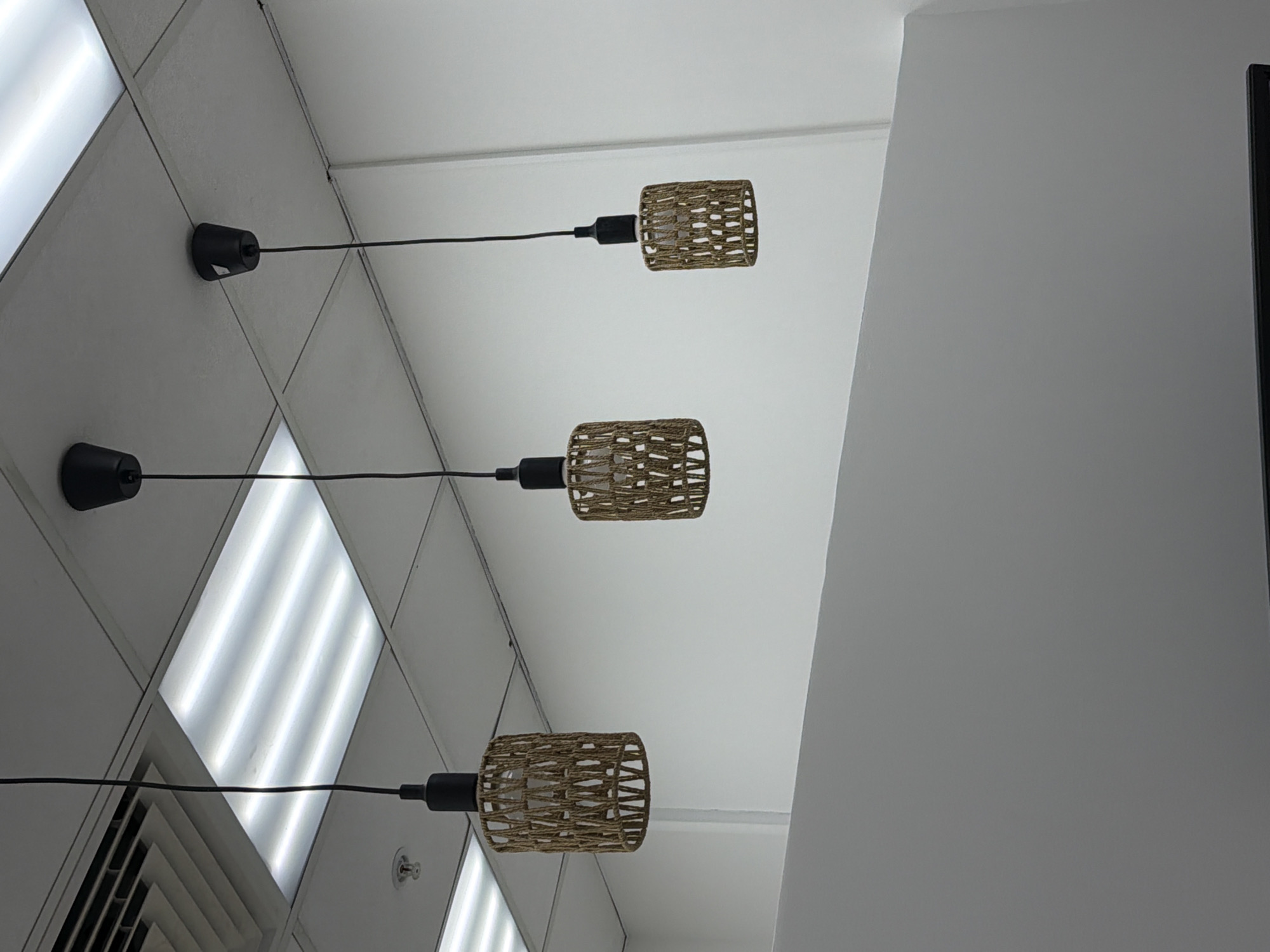 The image shows a low-angle view of a ceiling with three identical pendant lights. The ceiling appears to be composed of square panels, some of which contain recessed fluorescent lights. The pendant lights have black cords and cylindrical lamp shades with a woven, textured appearance, possibly made of natural fibers. A large, light gray wall is visible on the right side of the frame. The image provides no information to determine the location in Moscow, Russia, or any details about people, subjects, or actions.