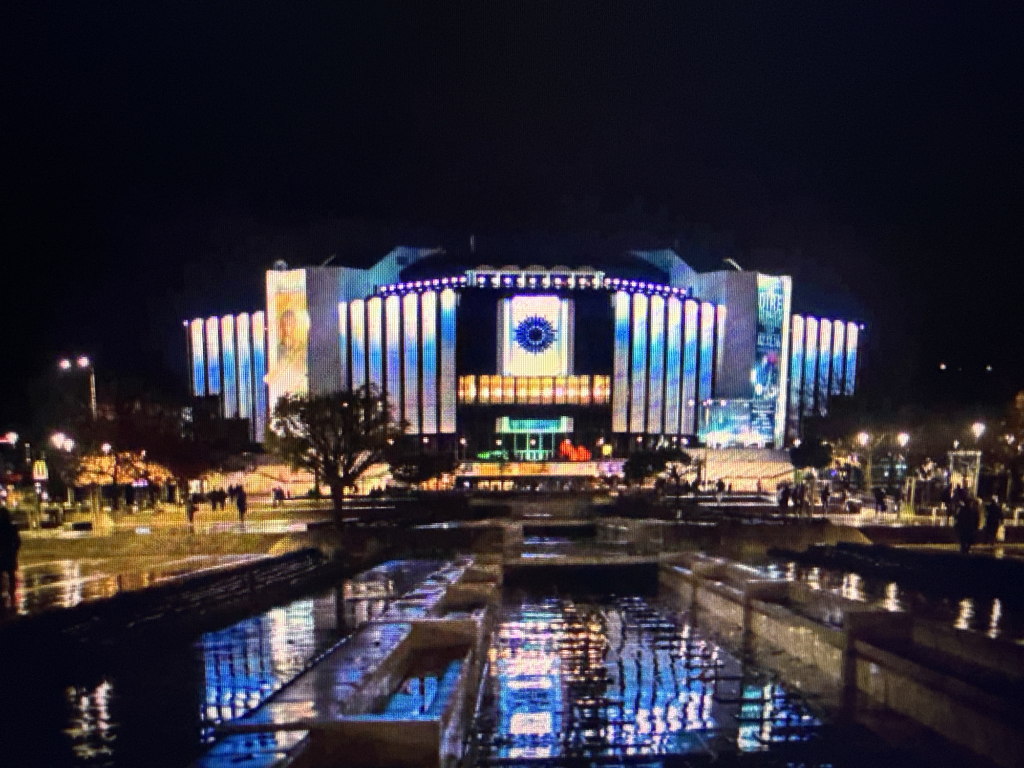 The image shows the National Palace of Culture (NDK) in Sofia, Bulgaria, illuminated with blue and white lights at night. The reflection of the building and lights is visible in the water feature in front of it. Pedestrians can be seen walking around the area. A tree is present in the foreground, partially obscuring the view of the NDK. The sky is dark, indicating it is nighttime.