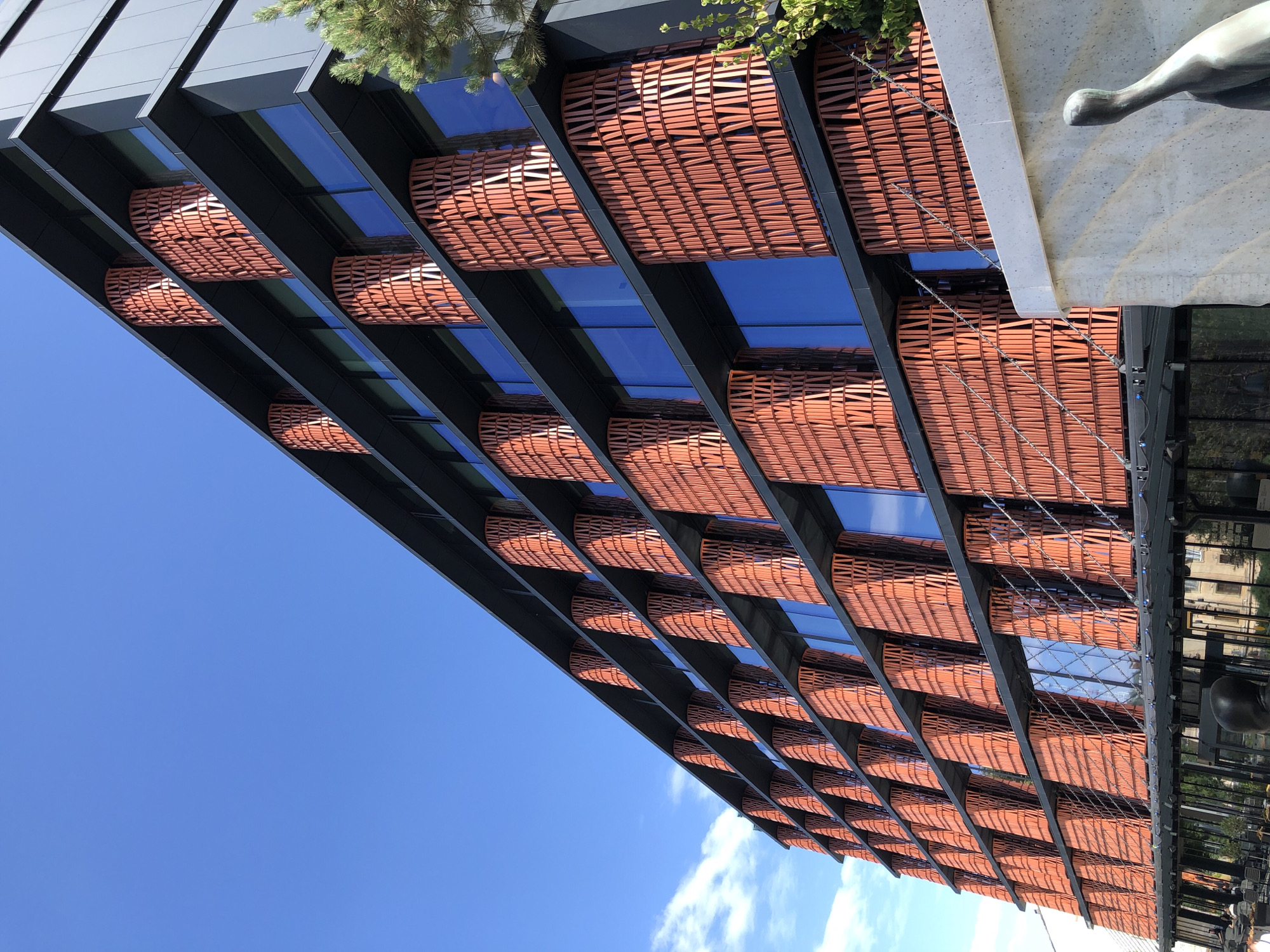 The image shows a modern building with a unique facade featuring a repeating pattern of orange, curved elements that appear to be made of terracotta or a similar material. These elements are arranged in vertical columns between rows of blue-tinted windows. The building is seen from a low angle, looking up towards a clear blue sky with some wispy clouds. To the right of the building, a portion of a green tree is visible, and in the foreground on the right, a light-colored concrete wall and a dark sculpture of a bird-like figure are present. The lower part of the building reveals a ground floor with glass windows and some outdoor seating areas with chairs and tables, suggesting it might be a commercial establishment like a restaurant or cafe. The architecture is contemporary and striking, with a strong emphasis on texture and geometric patterns. This building is the "Lviv House" located in Lviv, Ukraine.