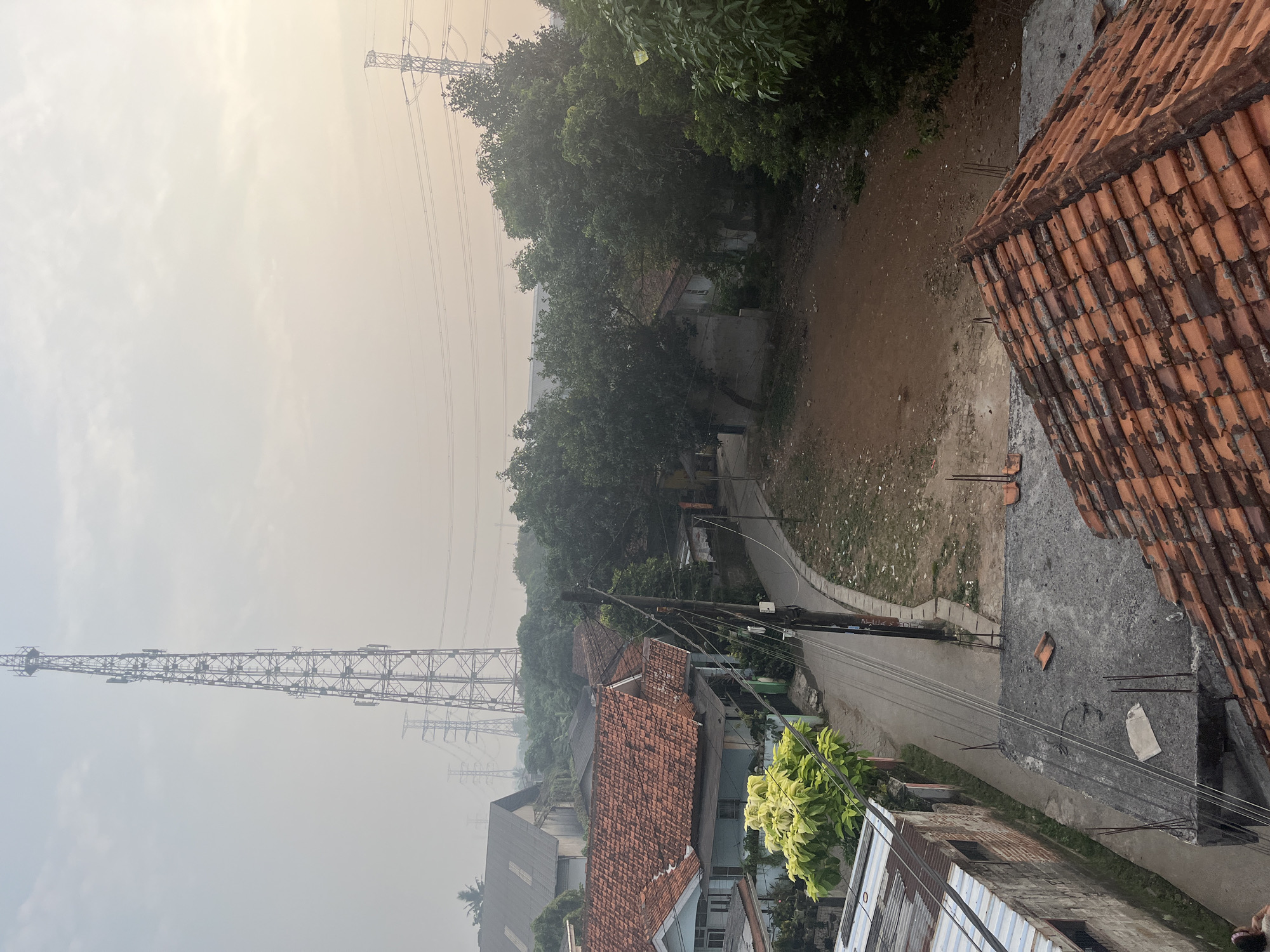The image depicts a hazy, overcast day in Pasir Gadung, Indonesia. The scene is dominated by a tall telecommunications tower in the distance, partially obscured by the atmospheric conditions. In the foreground, a residential area with red-tiled roofs is visible, along with a narrow road and surrounding vegetation. Power lines crisscross the sky, and electrical pylons can be seen in the mid-ground. The overall impression is one of a somewhat undeveloped urban or semi-urban environment. No people or specific actions are clearly identifiable in the image.