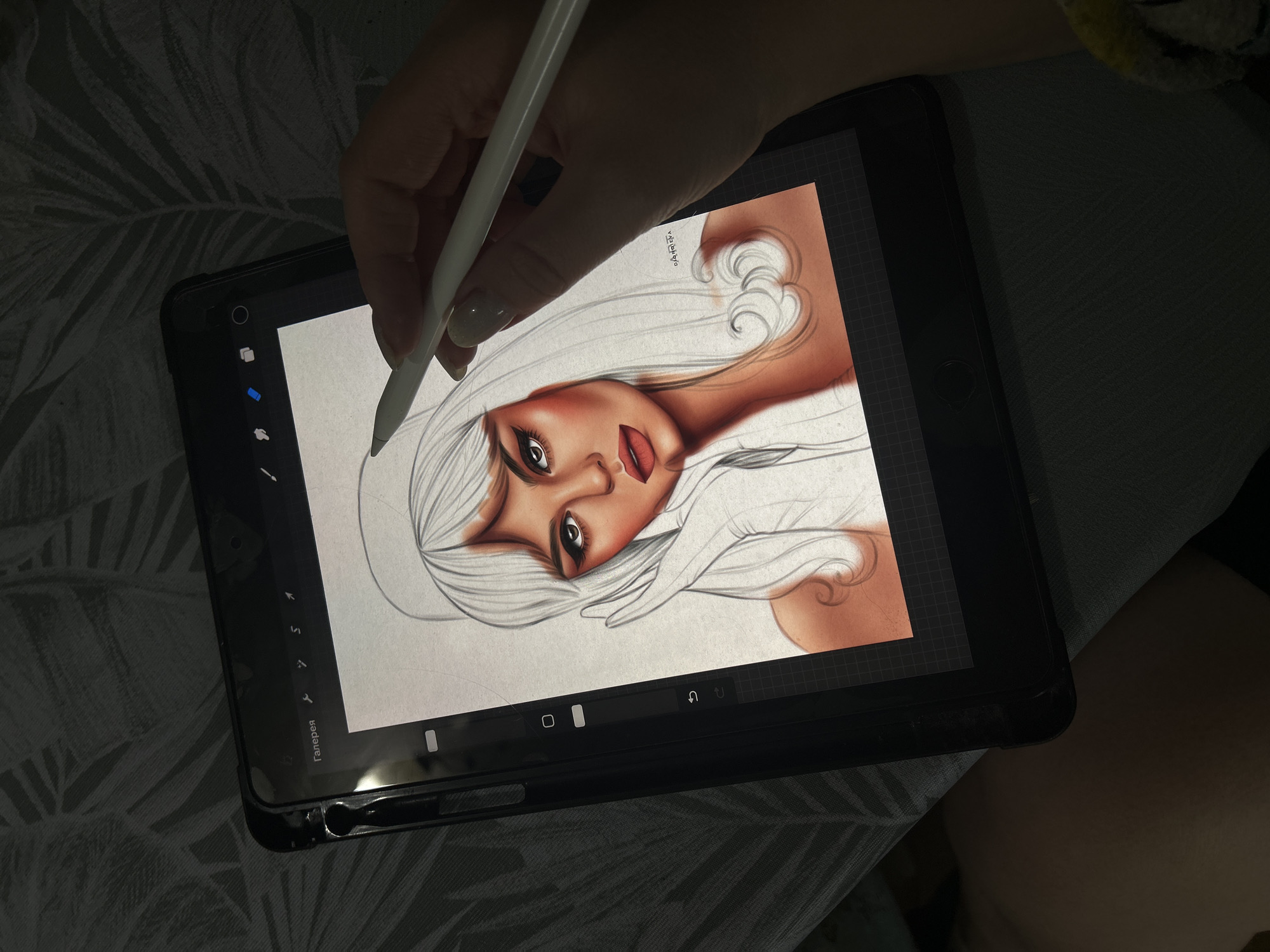 A hand holds a white stylus, drawing on a black tablet device displaying a partly finished digital portrait of a woman with pale skin, red lips, and white hair. The tablet is positioned against a patterned textile surface, possibly a blanket or bedding. The angle of the photograph is top-down, slightly tilted. The visible interface of the tablet shows digital drawing software with various tools displayed on the side. The portrait is predominantly in shades of white, red, and brown, appearing to be a work in progress. There is no indication that the location is Фоки, Russia.