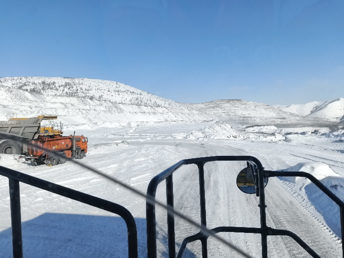 The image depicts an open-pit mining site in a snow-covered, mountainous environment, located in the city of Im. Gastello, Russia. The perspective is from inside a vehicle, with a dark metallic frame and a side-view mirror visible in the foreground. A dark shadow from the vehicle is cast on the snow-covered ground to the left. A wide, snow-covered road or path, marked with tire tracks, extends into the midground. To the left, a large, orange, multi-axle mining dump truck with a raised gray bed is stationary on the snow, with a yellow piece of heavy equipment visible behind it. The background consists of extensive snow-covered hills and mountains under a clear, bright blue sky. Several hills, particularly in the center and left-background, display terraced formations indicative of mining activity. Sparse dark vegetation is visible on some of the snow-covered slopes. No individuals are present, and no actions or interactions are discernible in the static scene.