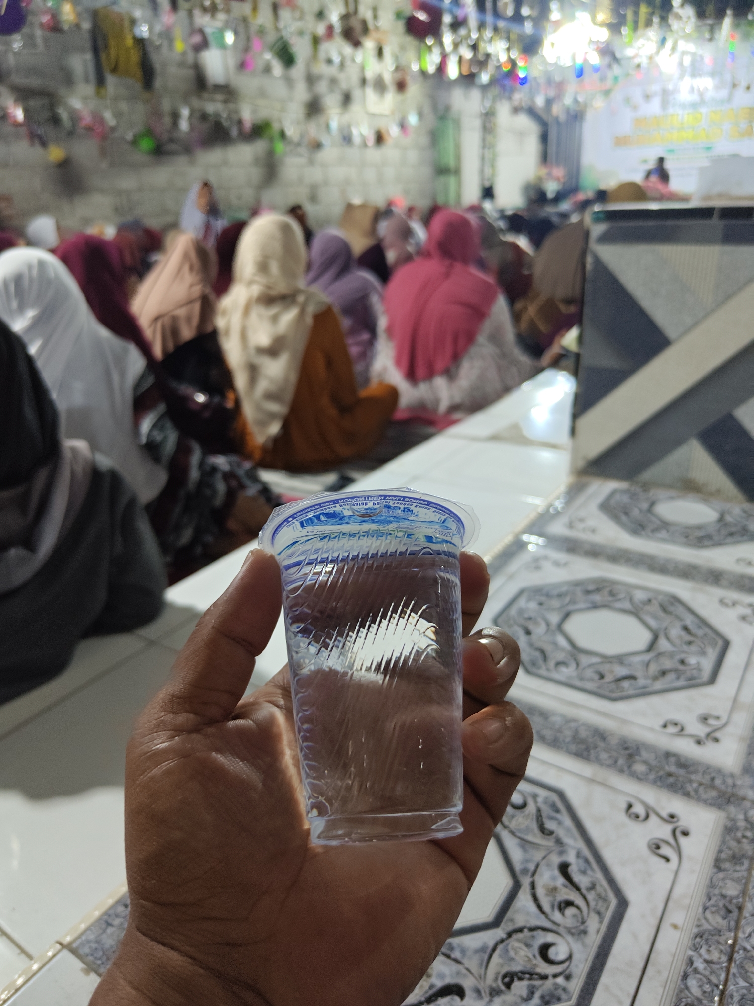 In Klampokan, Indonesia, a person's hand is holding a small, clear plastic cup filled with water. The cup has a blue and white label with text. The background is blurred but appears to be an indoor gathering, likely a religious event, with many people sitting on the floor, many wearing headscarves. The floor is tiled with a decorative pattern of octagons and swirling designs. To the right, there is a section of wall or partition with a geometric pattern of gray and white diagonal lines. The lighting in the room appears to be a mix of overhead artificial lights, some of which are colorful.